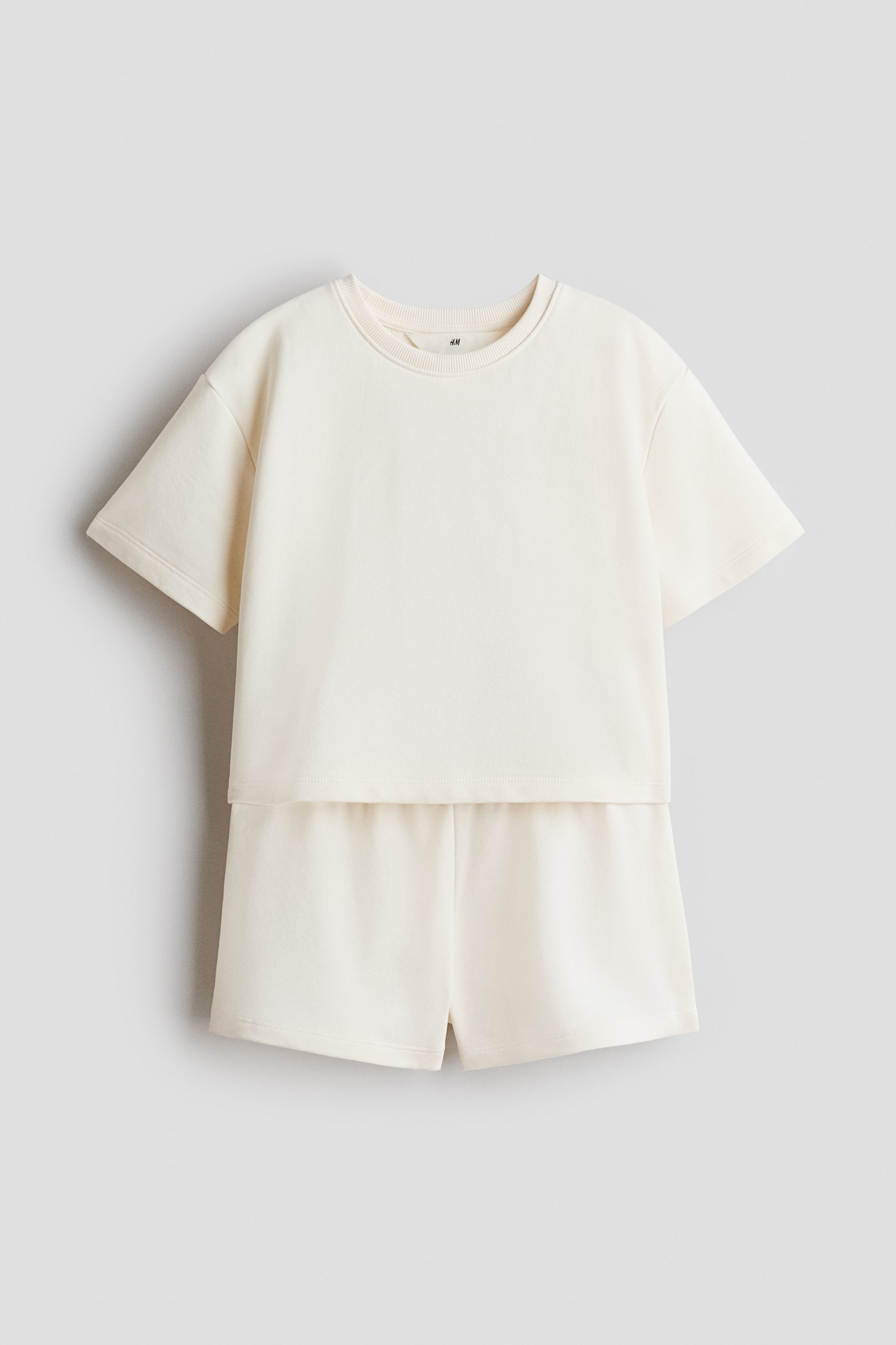 View larger image: A cream-coloured comfy set, crafted from lightweight sweatshirt fabric, is displayed flat and facing front. The t-shirt showcases a round, rib-trimmed neckline, dropped shoulders, and a straight-cut hem with short side slits, while the matching shorts are designed with covered elastication at the waist.