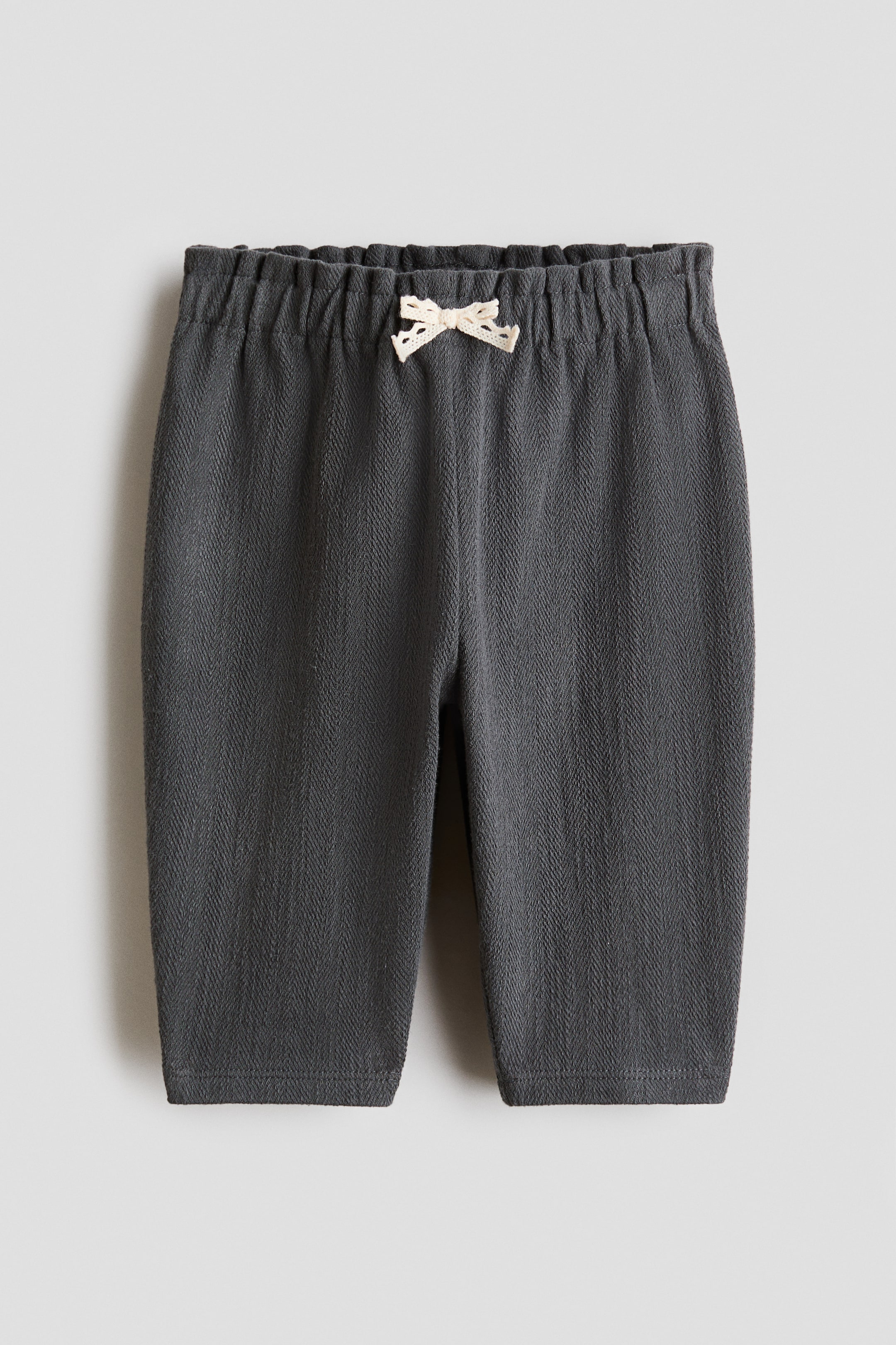 Ampliar la imagen: El frente de los pantalones gris oscuro muestra una tela con tejido de espiga. Una pretina elástica y fruncida presenta un pequeño lazo de encaje blanco roto atado en su centro.
