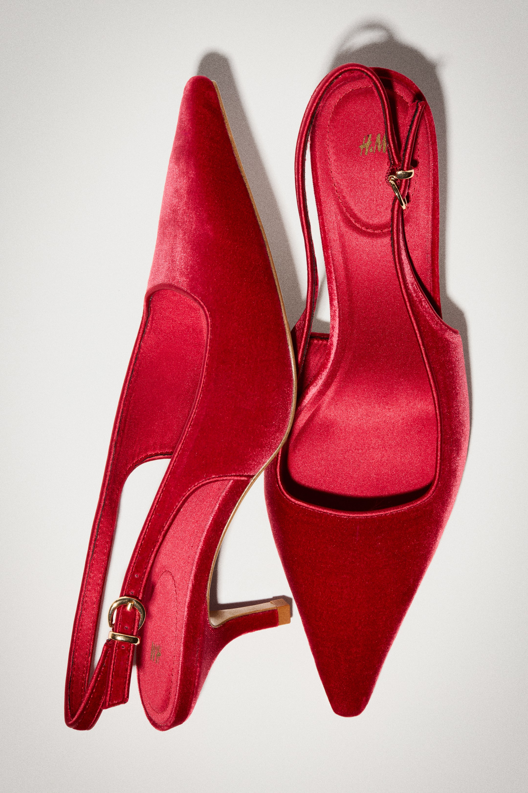 View larger image: A pair of red velvet slingback heels is lying flat, with one shoe shown in side profile to highlight the pointed toe, low stiletto heel, and narrow adjustable strap with a gold buckle. The other shoe is facing upward, showcasing the pointed toe and matching red satin lining and insole.