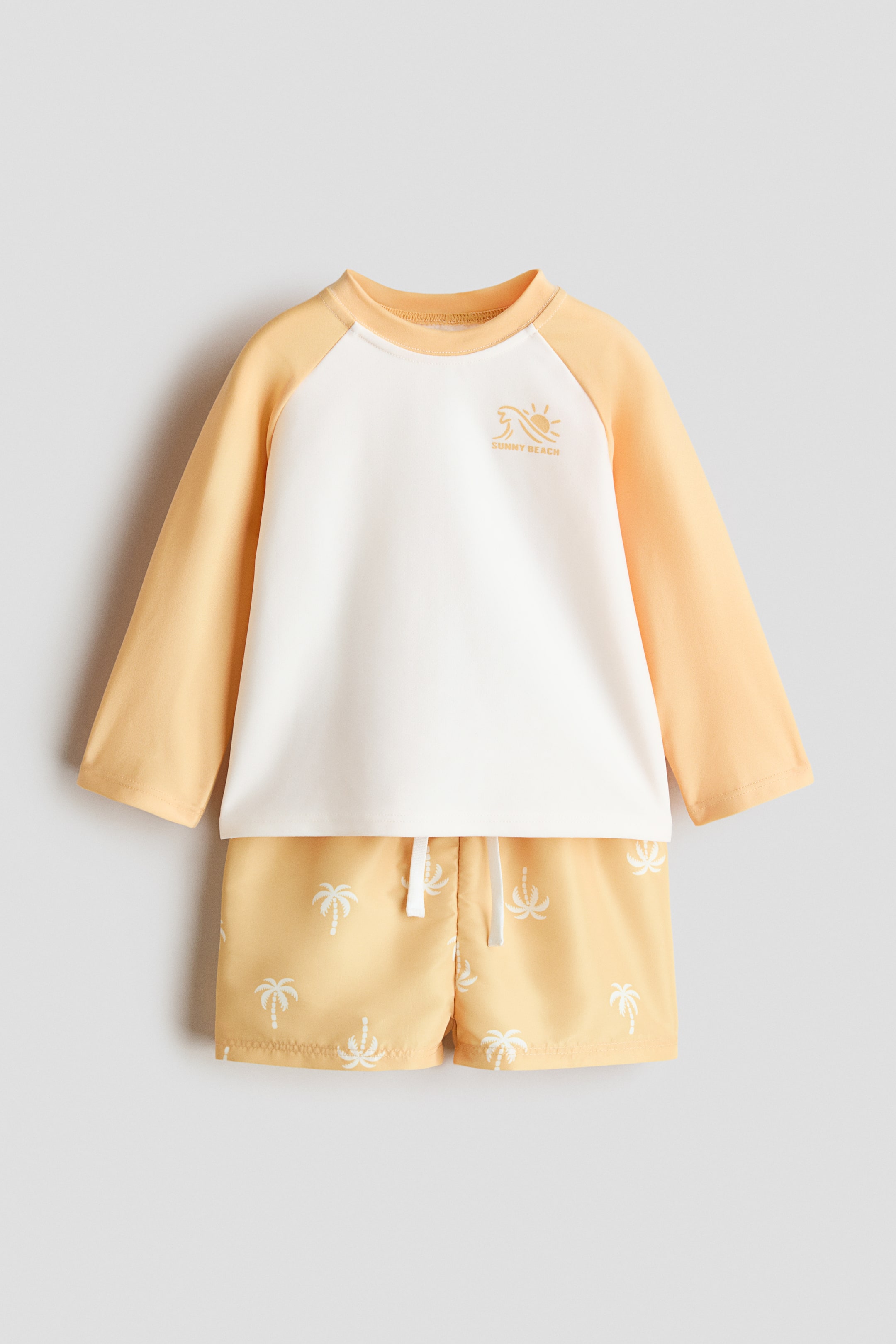 View larger image: A printed swim set is showcasing a long-sleeved top with a white body and light orange raglan sleeves, featuring a round trimmed neckline and a "SUNNY BEACH" graphic on the chest. It is paired with light orange shorts, patterned with white palm trees and featuring an elasticated drawstring waist.