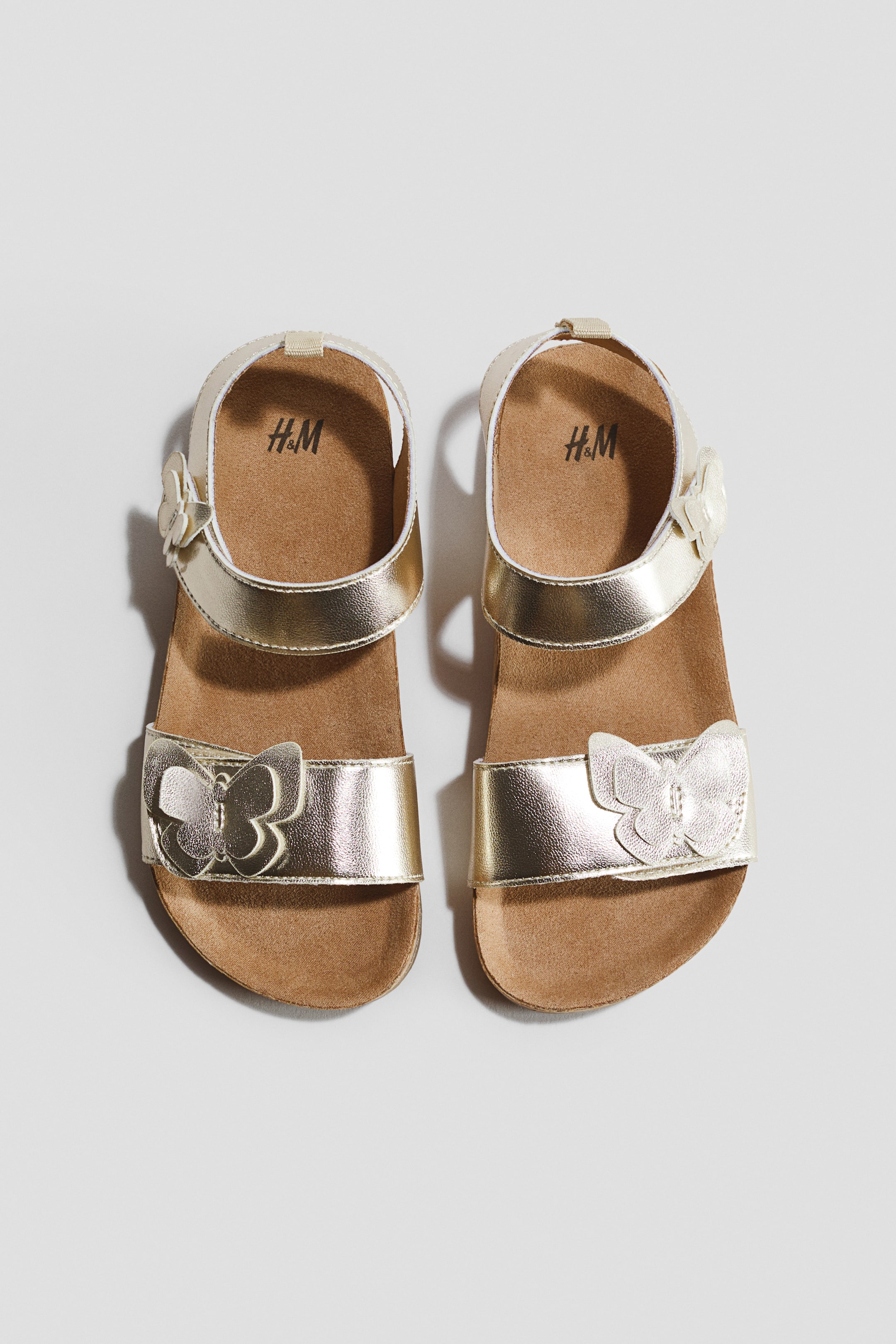 Visualizza immagine più grande: Un paio di sandali metallici dorati viene presentato dal FRONTE, caratterizzato da un cinturino sul piede e un cinturino alla caviglia, entrambi con chiusure regolabili a strappo e applicazioni a farfalla. I sandali presentano anche plantari sagomati e suole scanalate.