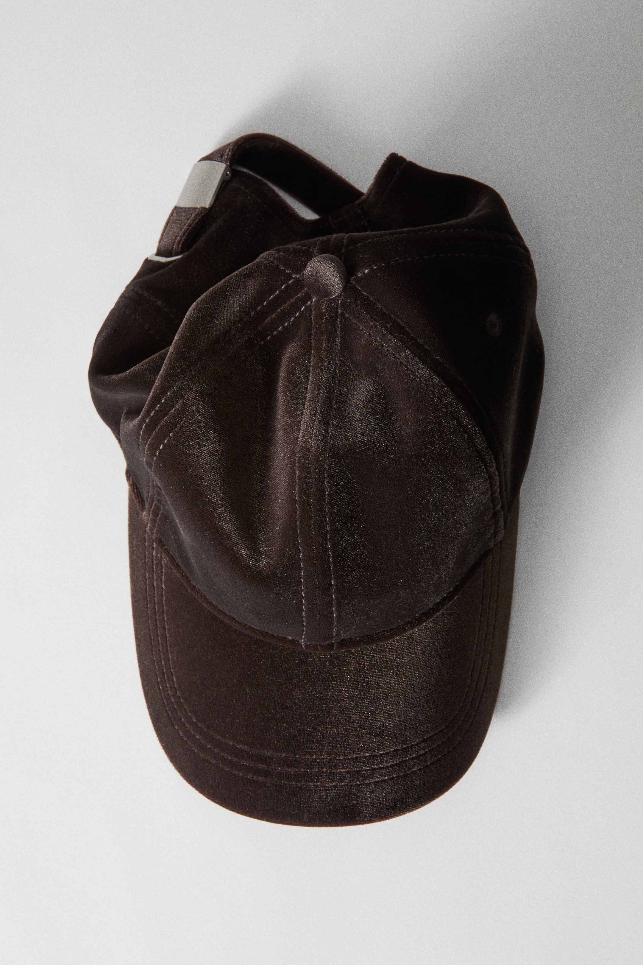 Ampliar la imagen: Se muestra una gorra de béisbol de terciopelo marrón oscuro desde un ángulo alto, exhibiendo su visera curva, la copa con paneles y botón superior, así como las costuras tonales. La parte trasera de la gorra es visible, con una tira ajustable de terciopelo y hebilla deslizante de metal plateado.