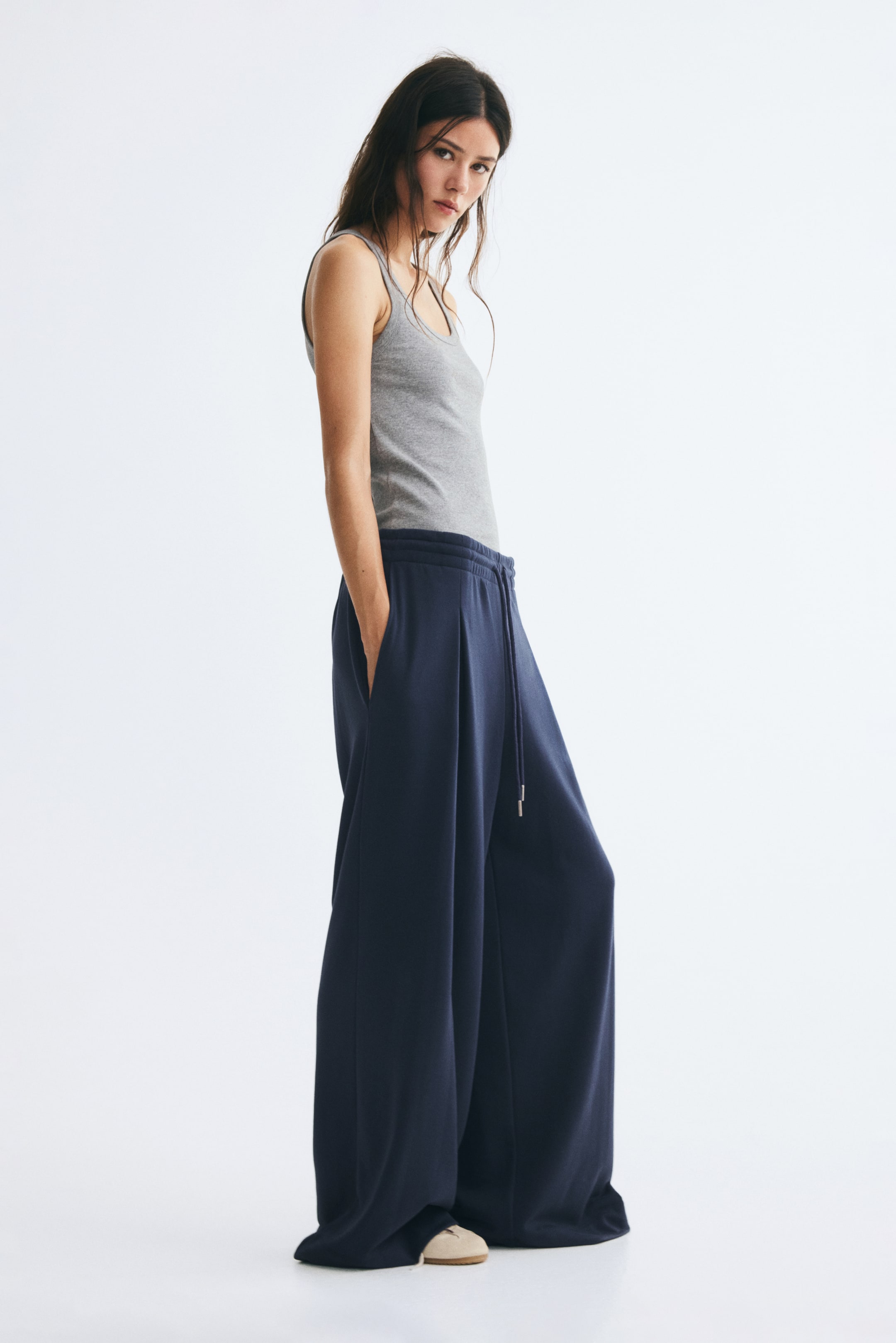 View larger image: Dark blue loose-fit pants in a lightweight sweat fabric are being worn by the model, featuring an elasticated drawstring waist, front inverted box pleats, and extra-long, wide legs. The pants are styled with a tucked-in grey tank top, with the model standing and holding one hand in the side pocket.