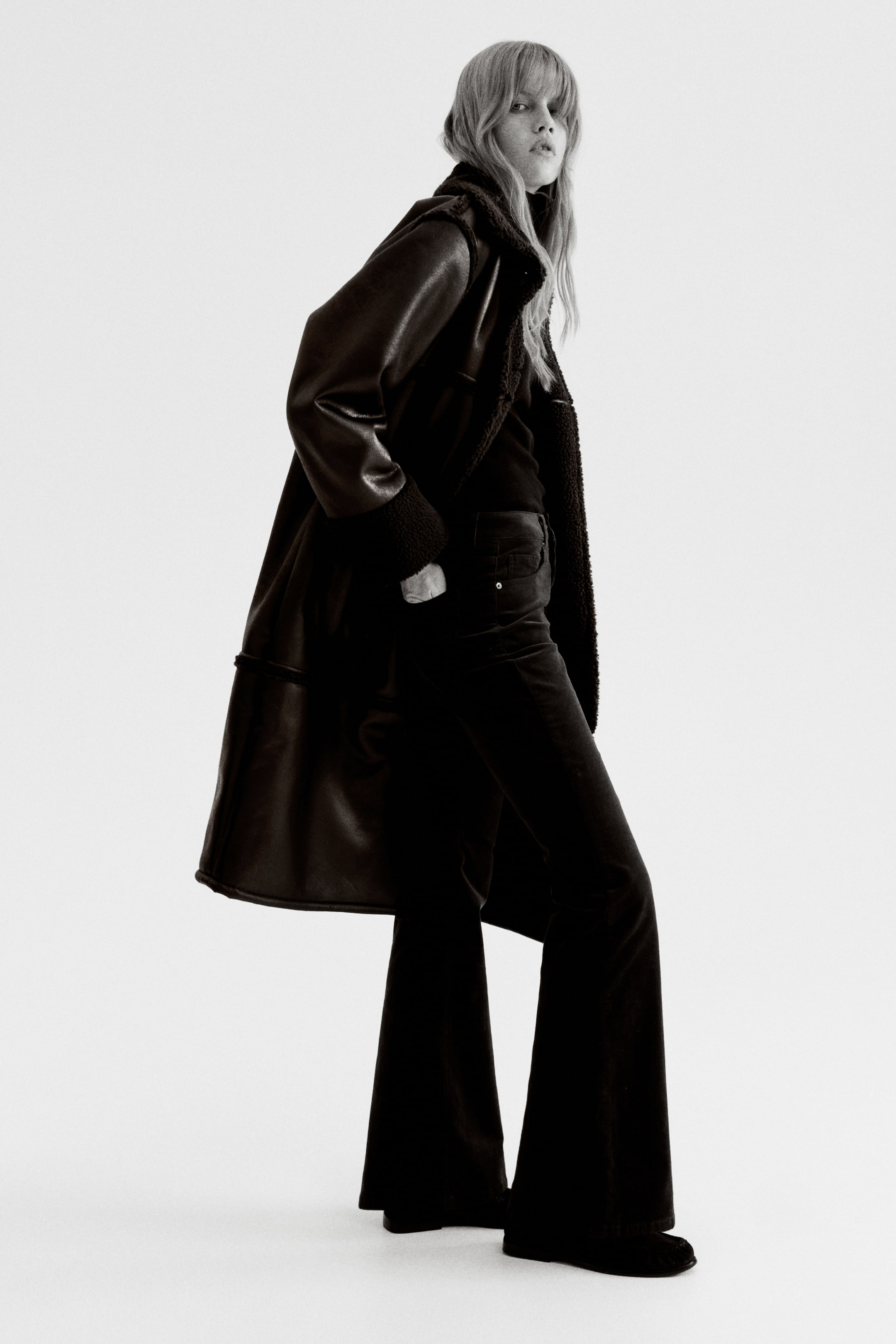 Größeres Bild anzeigen: Ein langer, dunkler Mantel, möglicherweise aus Kunstleder oder Wildleder, wird aus einem leichten rechten Winkel präsentiert. Er hat einen breiten Kragen und ein kontrastierendes flauschiges Futter, das am Kragen, an den Ärmelbündchen und an den inneren Nahtlinien sichtbar ist, getragen über dunklen, ausgestellten Hosen und einem dunklen Oberteil.