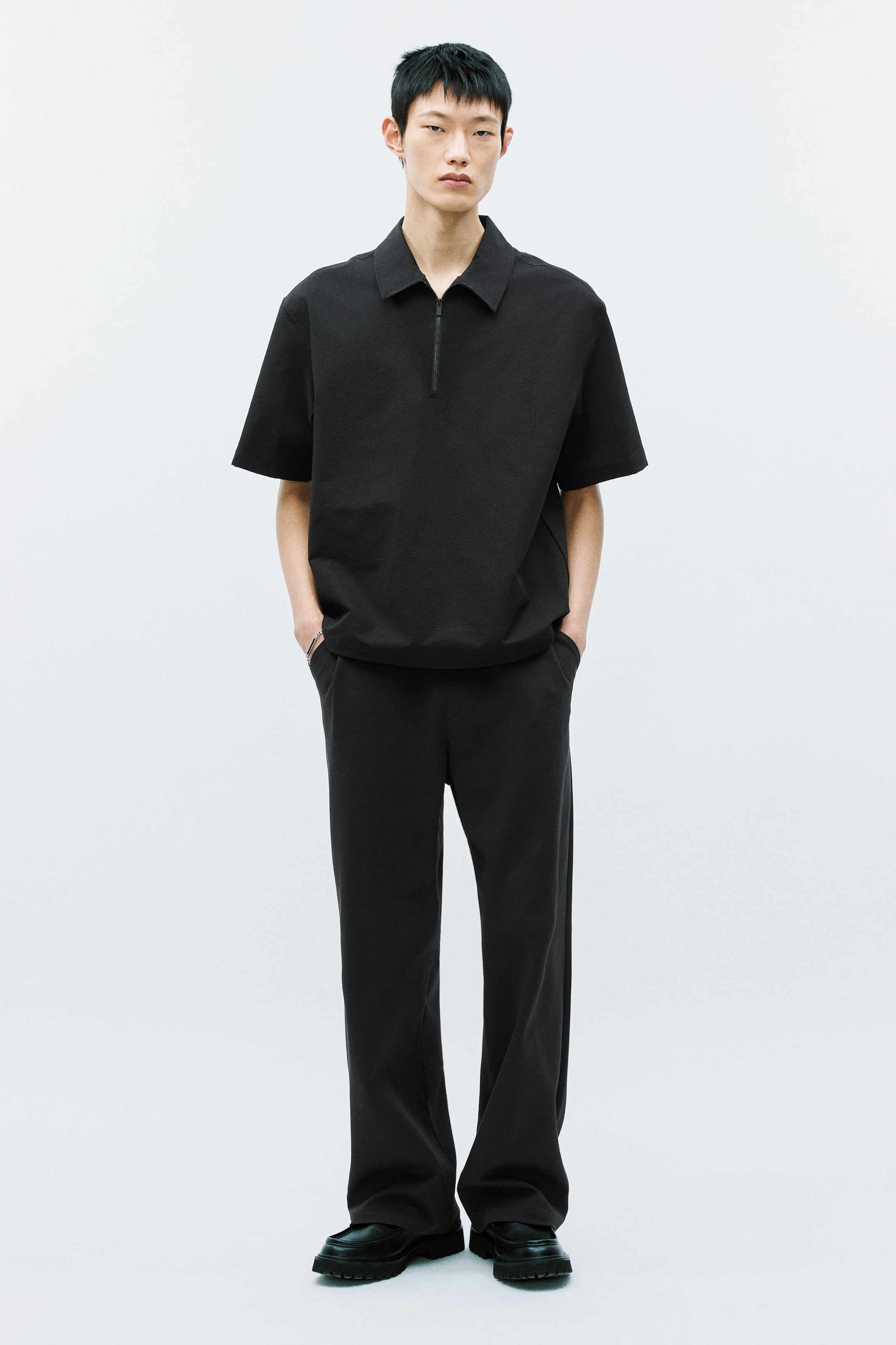 拡大画像を表示: 黒のテクスチャード加工された半袖ポロシャツは、ポイントカラー、ハーフジップクロージャー、ストレートヘムのリラックスフィットで着用されており、黒のストレートレッグパンツと黒の厚底ローファーと共に、全て前向きに配置されています。