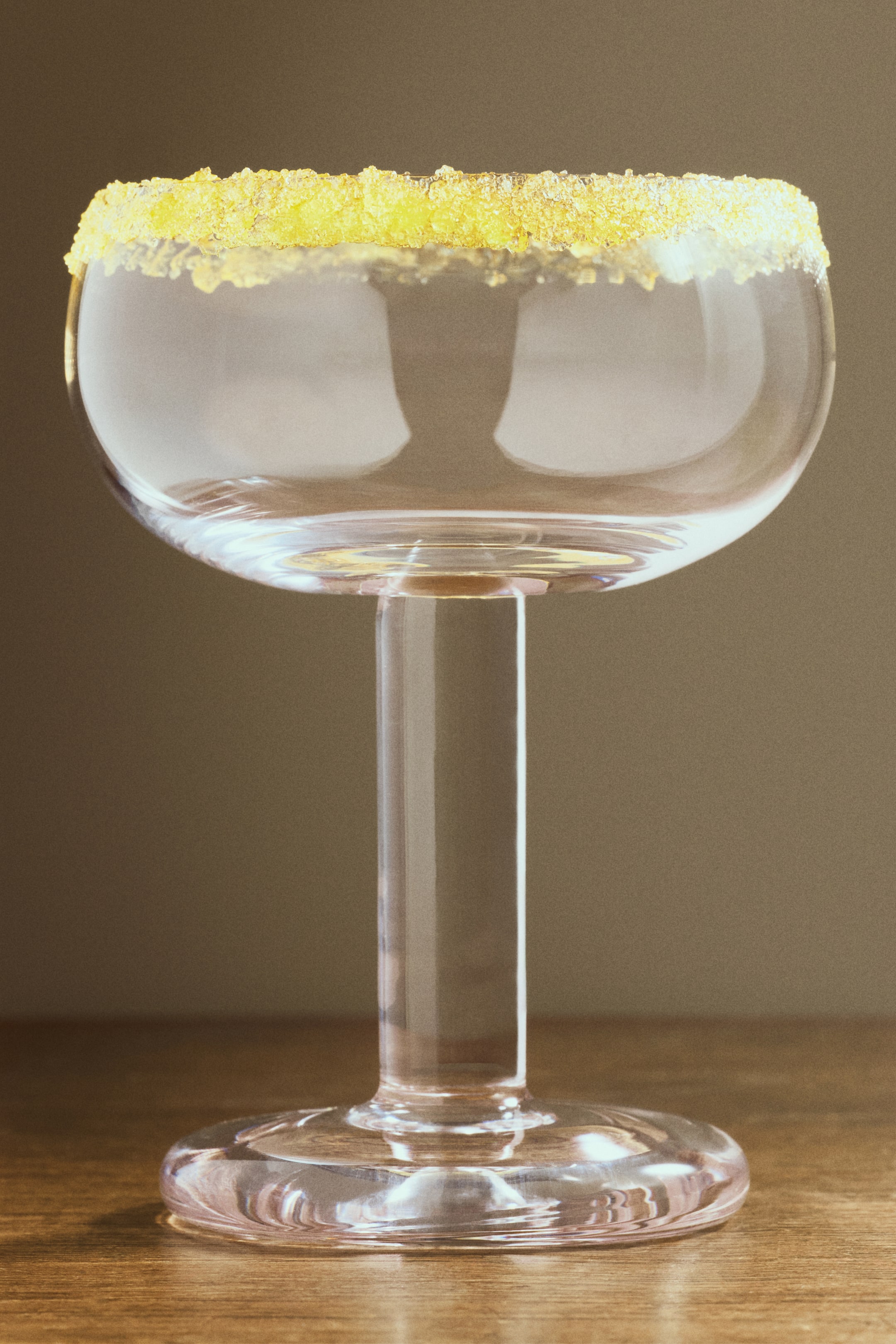 Voir une image plus grande: Un verre à coupe orienté vers l'avant présente un bol doucement arrondi, une tige audacieusement teintée subtilement, et une base circulaire. Le bord du verre est décoré de cristaux de sucre jaunes.