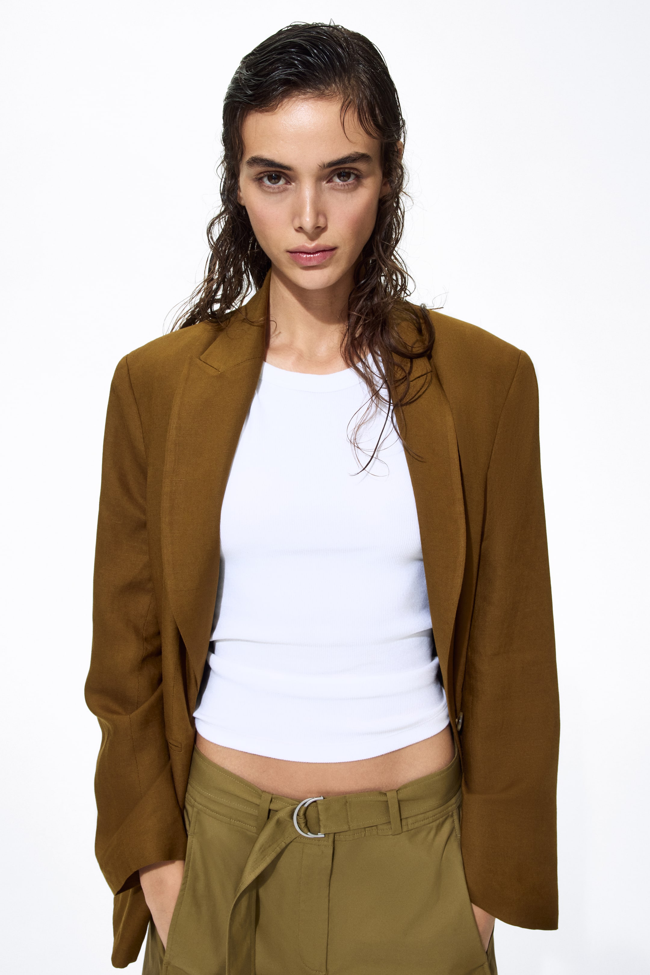View larger image: A structured brown blazer is worn open, showcasing a white ribbed tank top. It's paired with olive green trousers featuring a gathered waist and a silver D-ring belt.