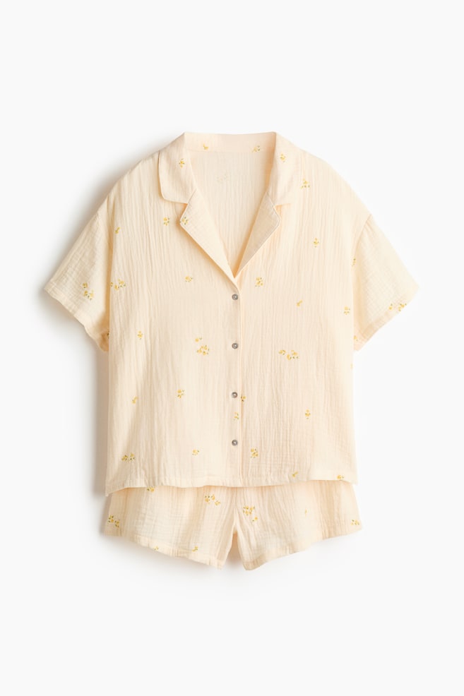 Ein hellgelbes Pyjama-Set zeigt ein kurzärmeliges Oberteil und passende Shorts, beide mit kleinen gelben Blumenstickereien gemustert. Das Oberteil verfügt über einen Kragen, eine durchgehende Knopfleiste und kurze Ärmel, wobei der Stoff geknittert erscheint. - 1