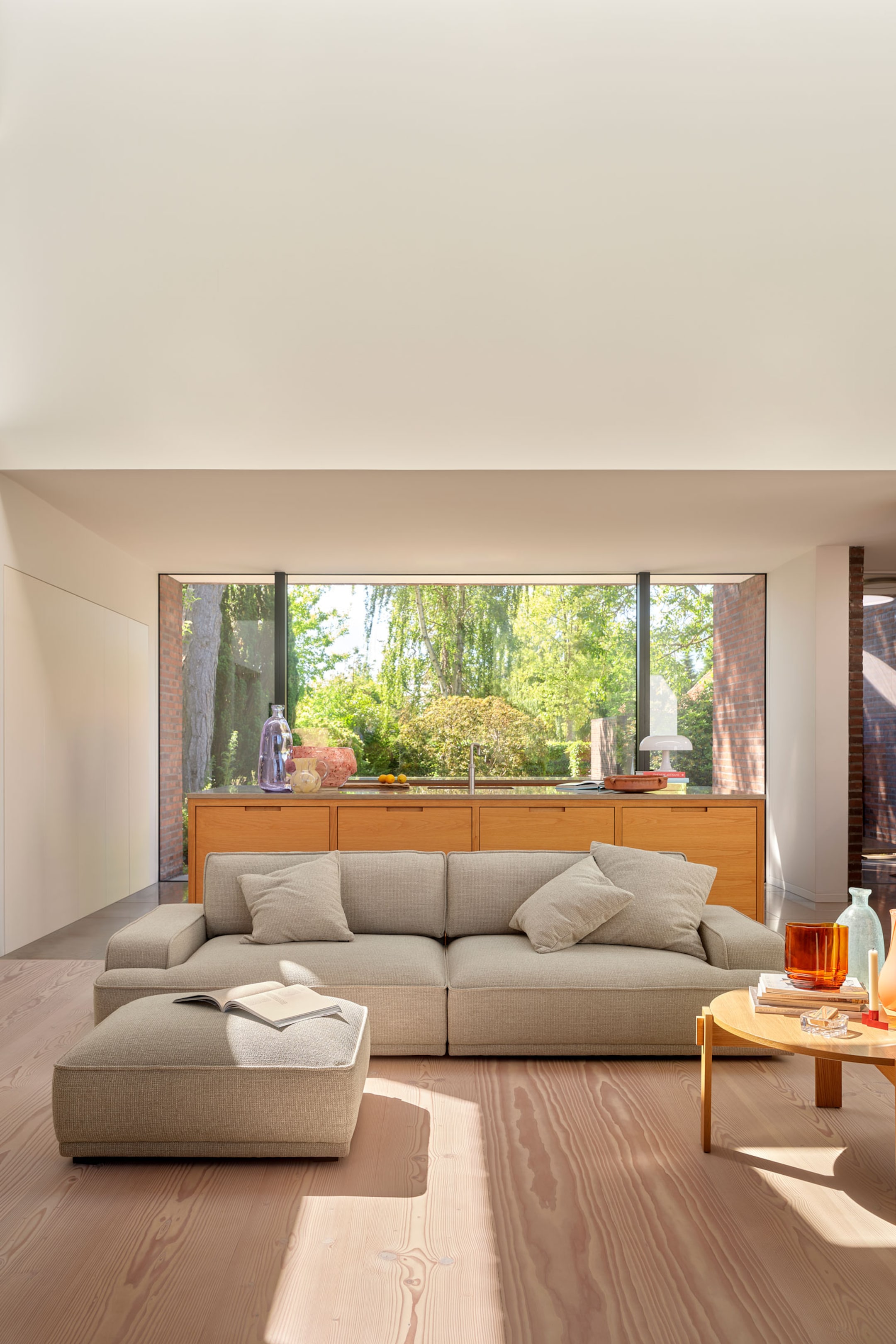 Größeres Bild ansehen: Ein niedriges modulares Sofa, bezogen mit einem hellbeigen, strukturierten Stoff, ist nach vorne ausgerichtet und verfügt über blockförmige Armlehnen, tiefe Polster und drei passende Zierkissen. Ein dazu passender quadratischer Ottoman im gleichen Stoff ist vor der linken Seite des Sofas positioniert.