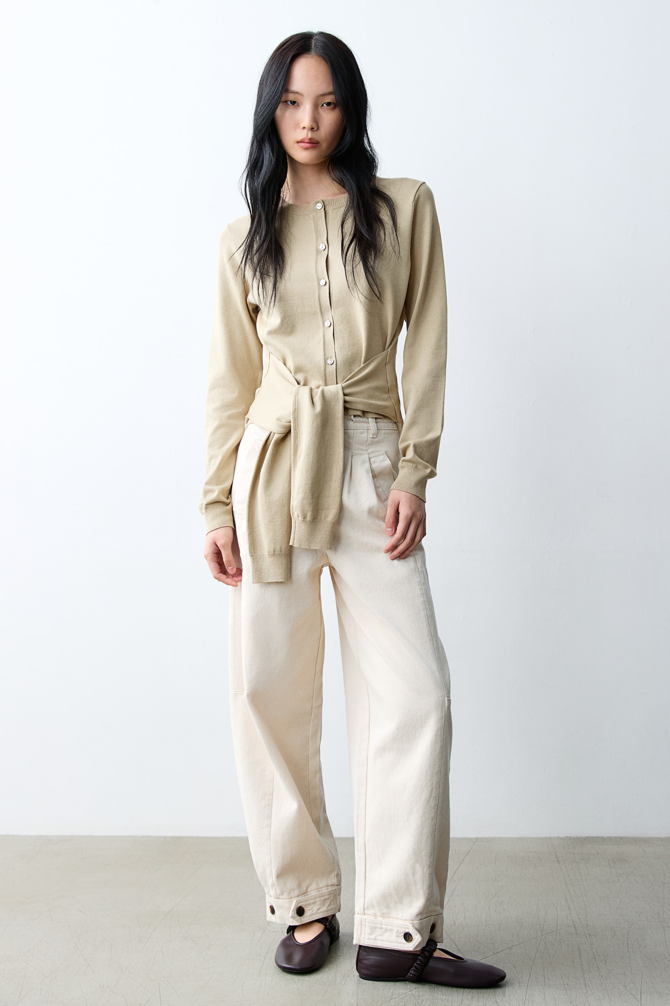 拡大画像を表示: ライトベージュのクルーネックカーディガンを着用しており、前面にボタン留めがあり、長袖です。同じカーディガンがウエストに結ばれています。ワイドレッグのプリーツクリームパンツも着用しており、裾には2つのボタンが付いたカフスが施されています。