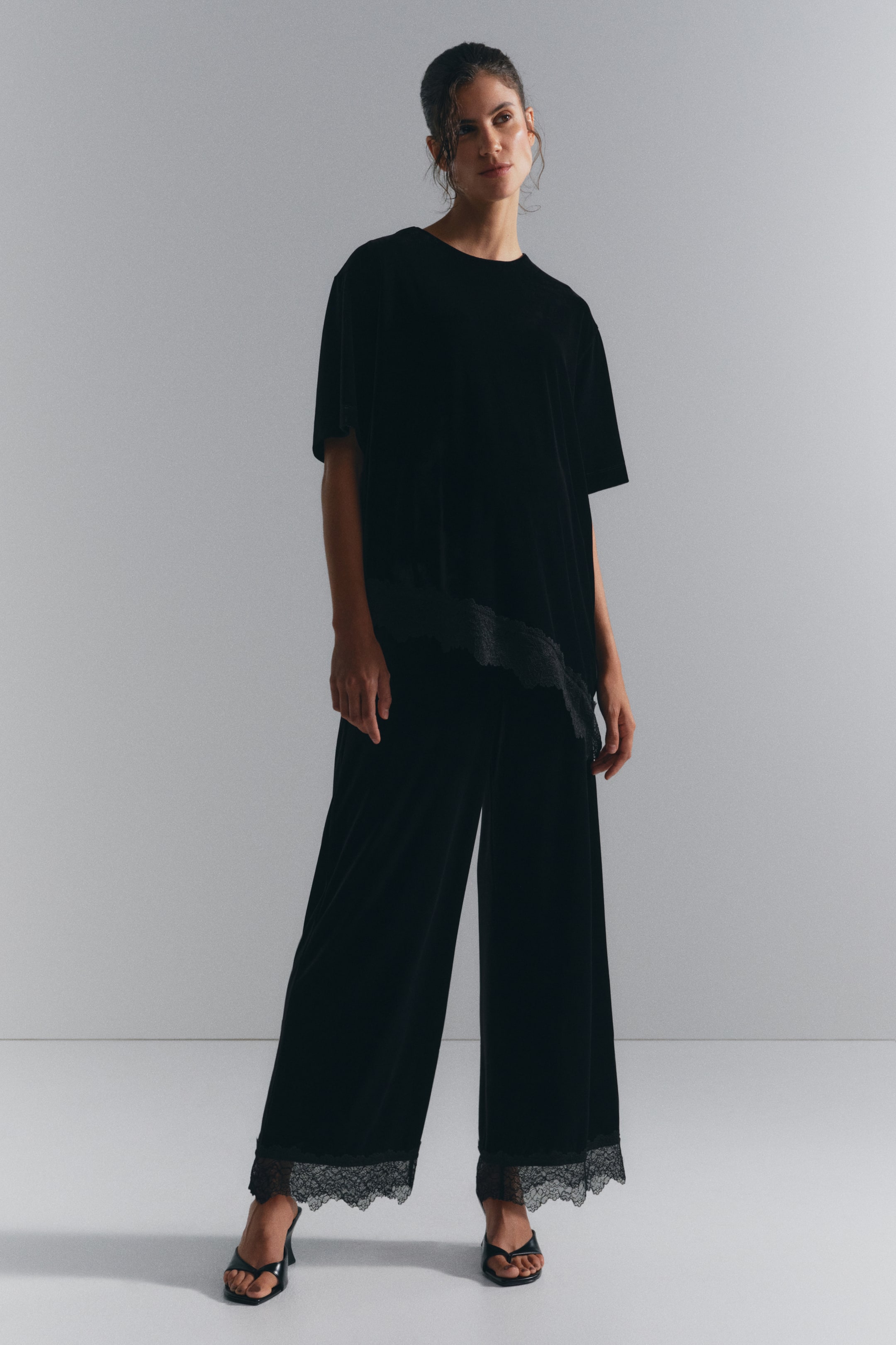 Προβολή μεγαλύτερης εικόνας: Ένα σετ loungewear από μαύρο βελούδο, που περιλαμβάνει ένα ασύμμετρο τοπ με κοντά μανίκια και ασορτί παντελόνι με φαρδιά μπατζάκια, φοριέται από το μοντέλο. Και τα δύο κομμάτια διαθέτουν λεπτεπίλεπτες μαύρες δαντελένιες λεπτομέρειες στα τελειώματά τους, ενώ το μοντέλο συνδυάζει το σύνολο με μαύρα πέδιλα με τακούνι.