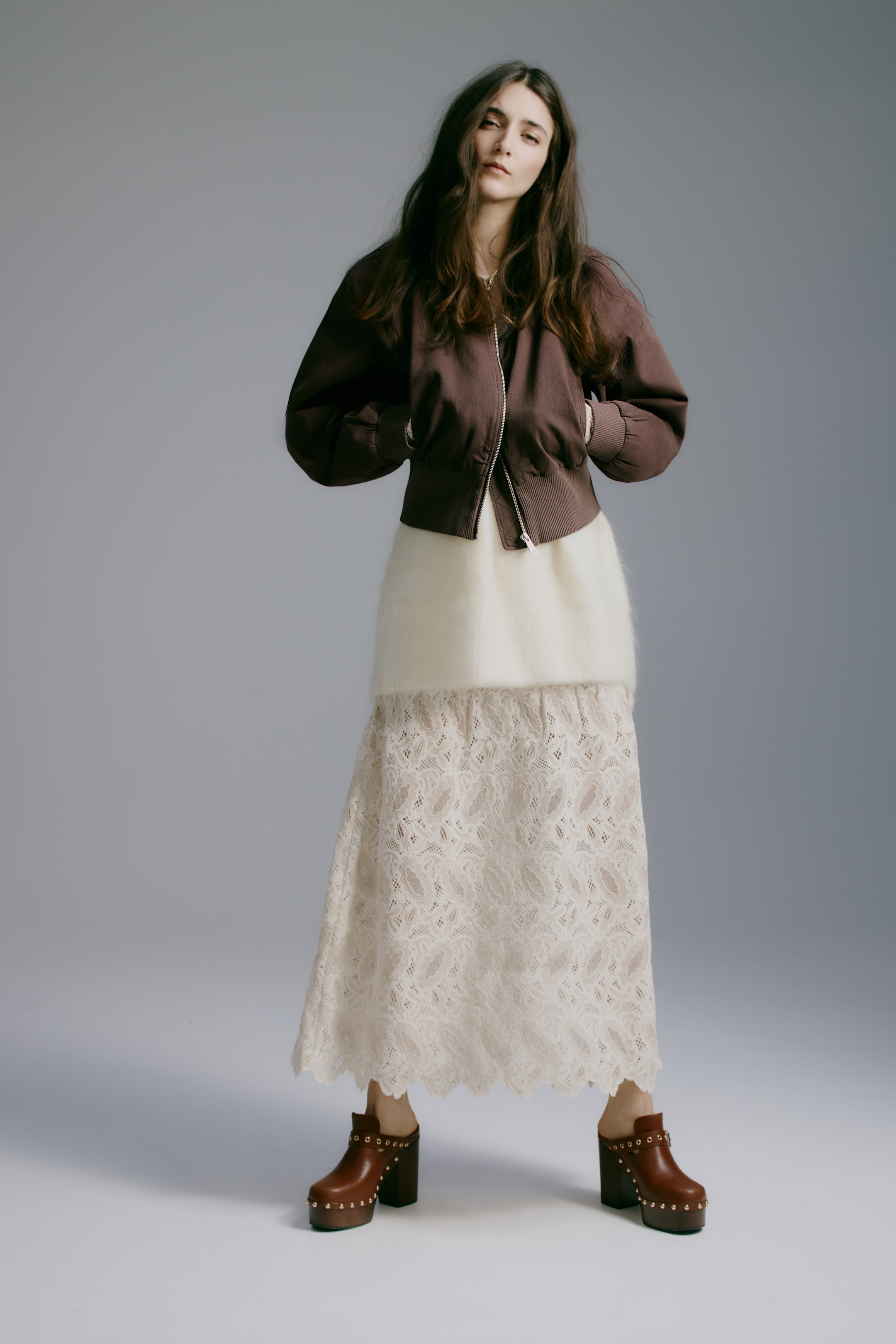 View larger image: A dark brown bomber jacket with ribbed cuffs and hem is showcased front-on, layered over a cream-colored fuzzy top and a long cream lace skirt with a scalloped hem. The ensemble is completed with brown platform clogs featuring studded details and wooden soles.