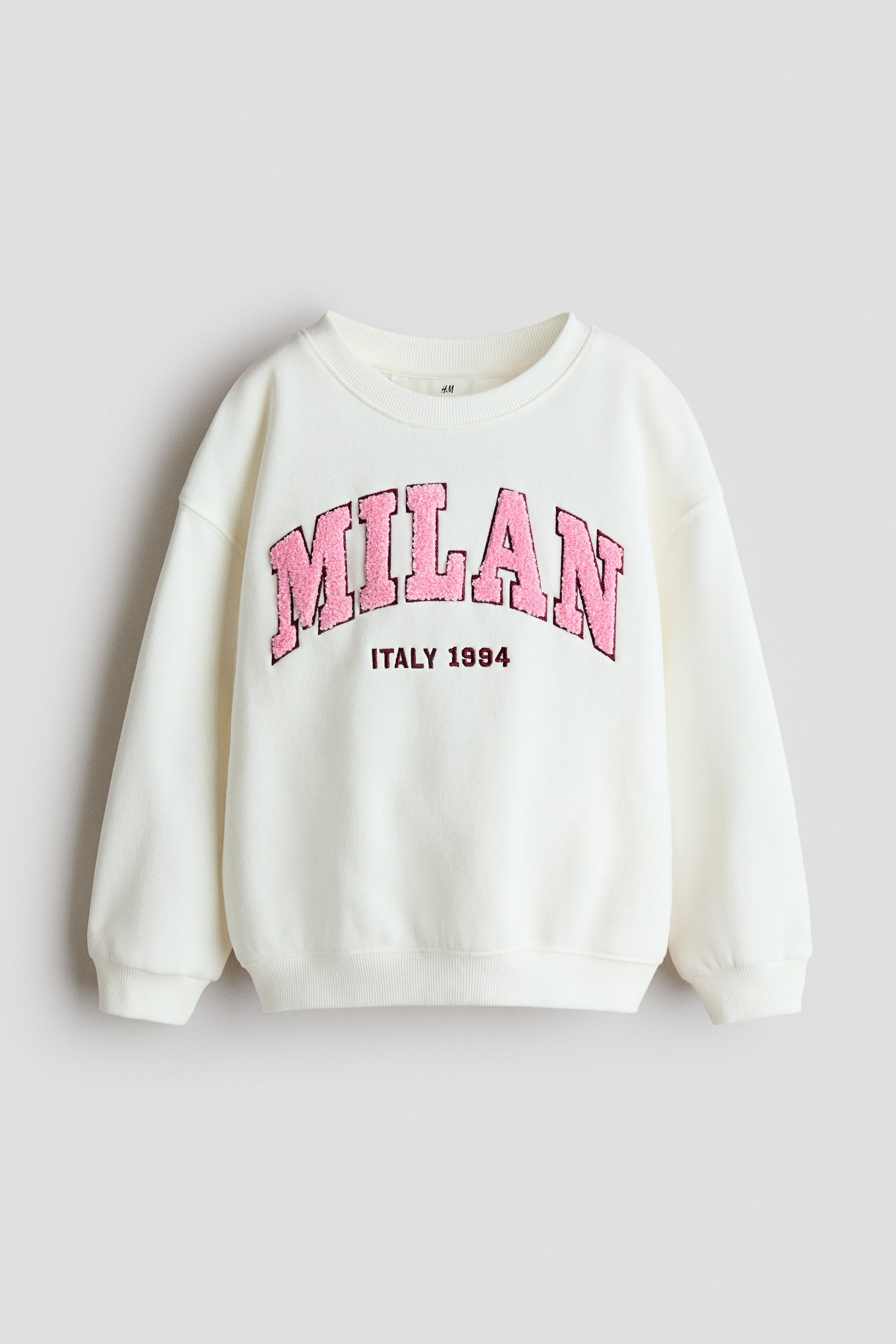 View larger image: An off-white sweatshirt showcases a crew neck, long sleeves with ribbed cuffs, and a ribbed hem. The front features the arched word 'MILAN' in large, pink, textured letters with a dark outline, and 'ITALY 1994' is printed below.