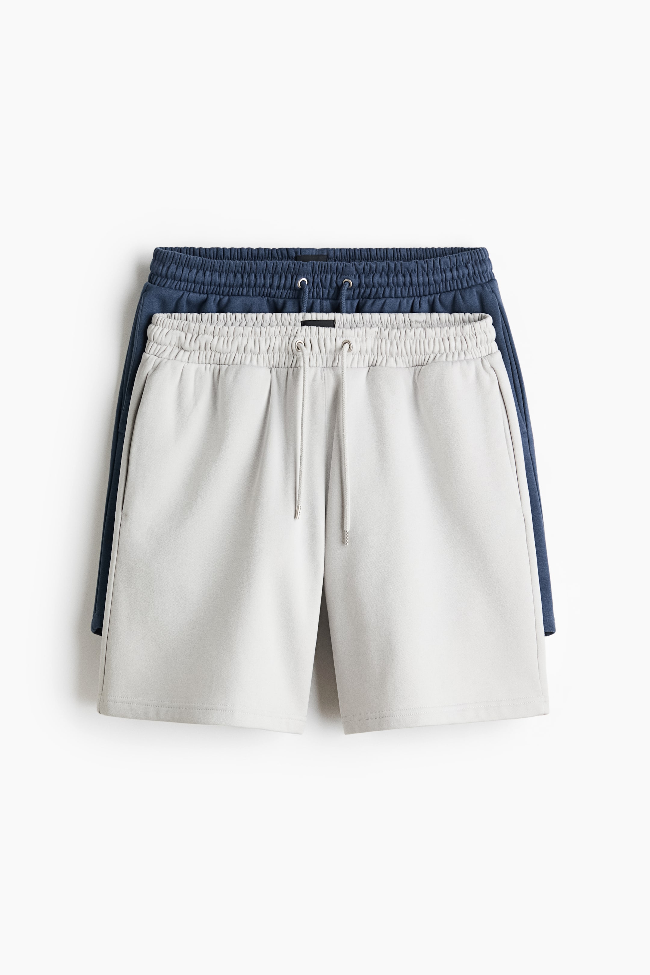 View larger image: Light gray sweatshirt shorts are lying flat and viewed from the front, featuring an elastic drawstring waist and side pockets, with a dark blue pair of shorts partially visible underneath.