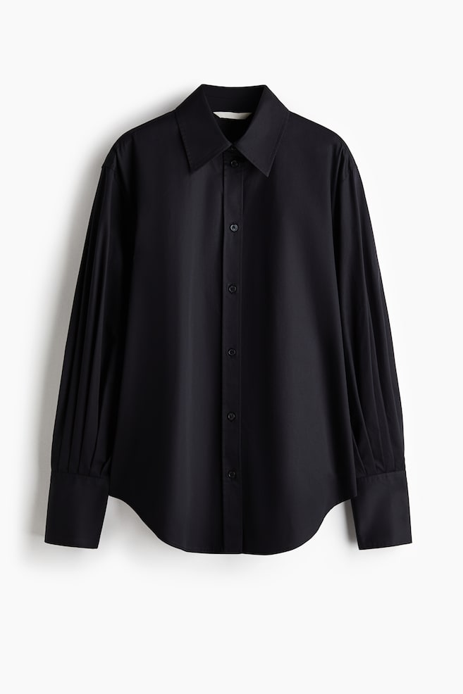 A front view of a black button-down shirt is showcasing a classic pointed collar, a button-front closure, and a curved hemline. The long sleeves are featuring distinctive vertical pleating that drapes down to wide, structured cuffs. - 1