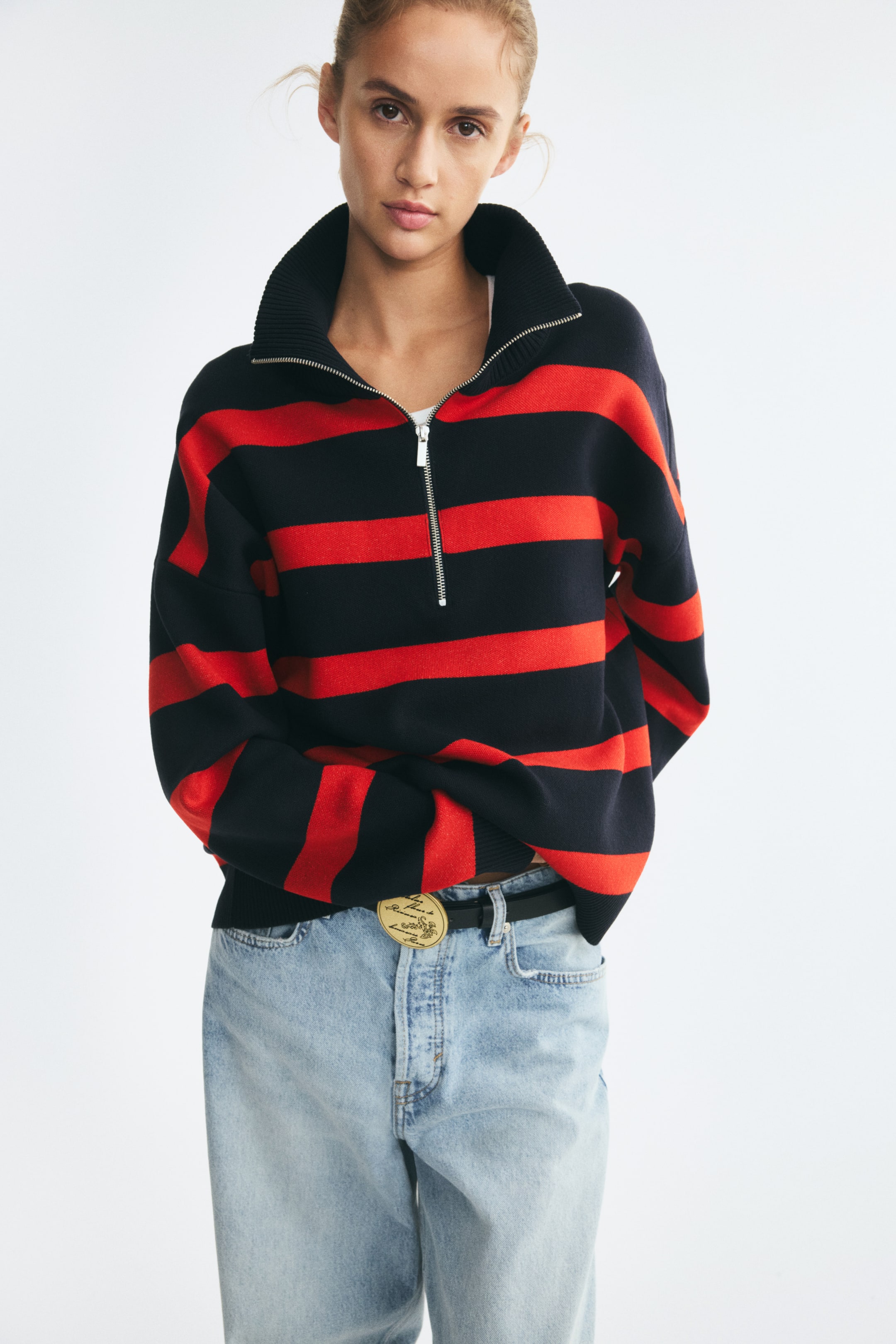 Agrandir l'image: Un pull en maille oversize à rayures horizontales noires et rouges, avec un col montant côtelé et une demi-fermeture zippée partiellement ouverte, est porté par le mannequin. Le pull présente des épaules tombantes et des poignets côtelés, tandis que le mannequin glisse une main sous l’ourlet pour mettre en valeur un jean délavé clair, porté avec une ceinture noire.