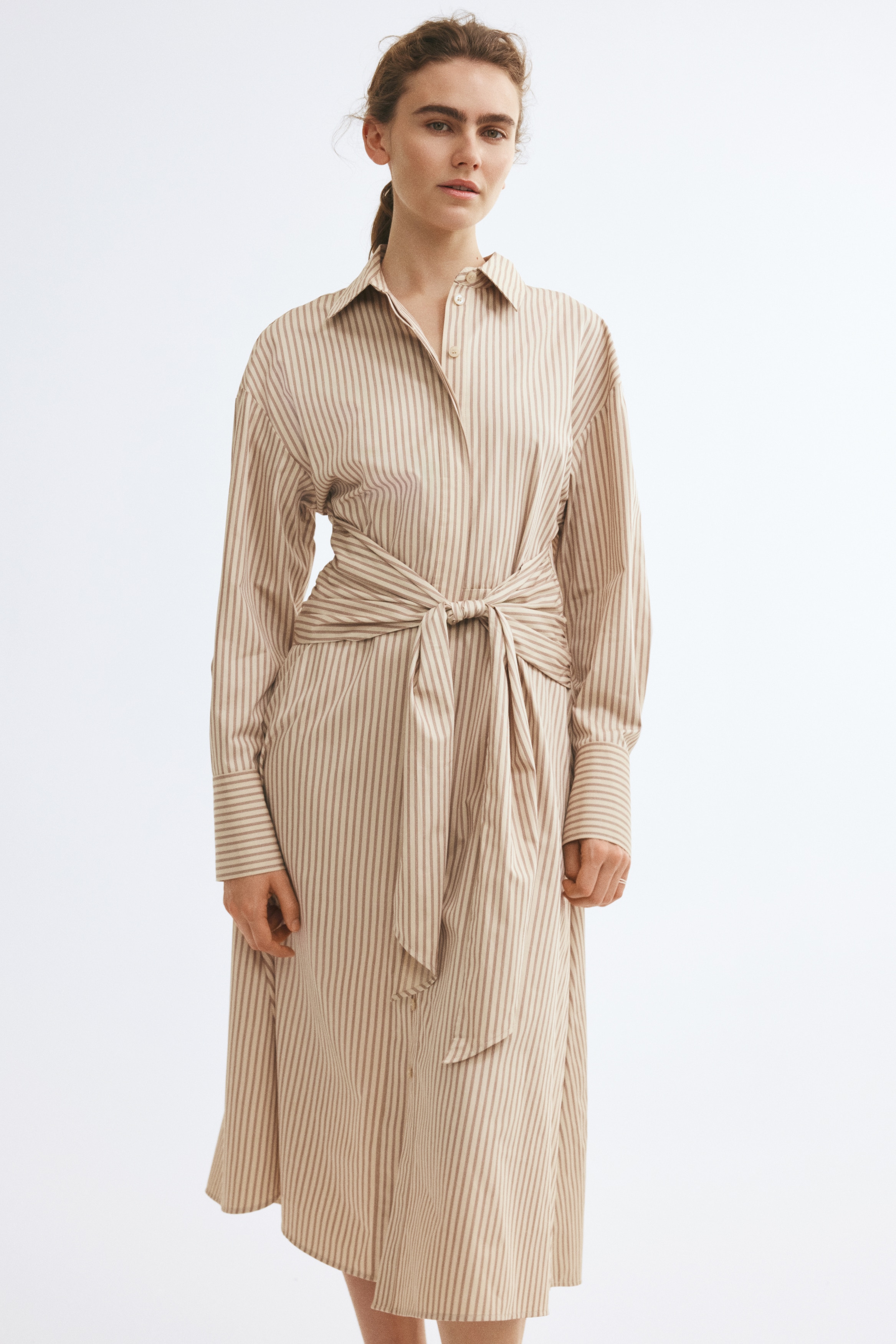 A calf-length shirt dress in a beige-and-white vertical stripe pattern is shown facing front, featuring a pointed collar, a concealed button placket, and dropped shoulders with long sleeves. The design is highlighted by draped panels on the sides of the bodice that tie into a large knot at the waist, cinching the silhouette.