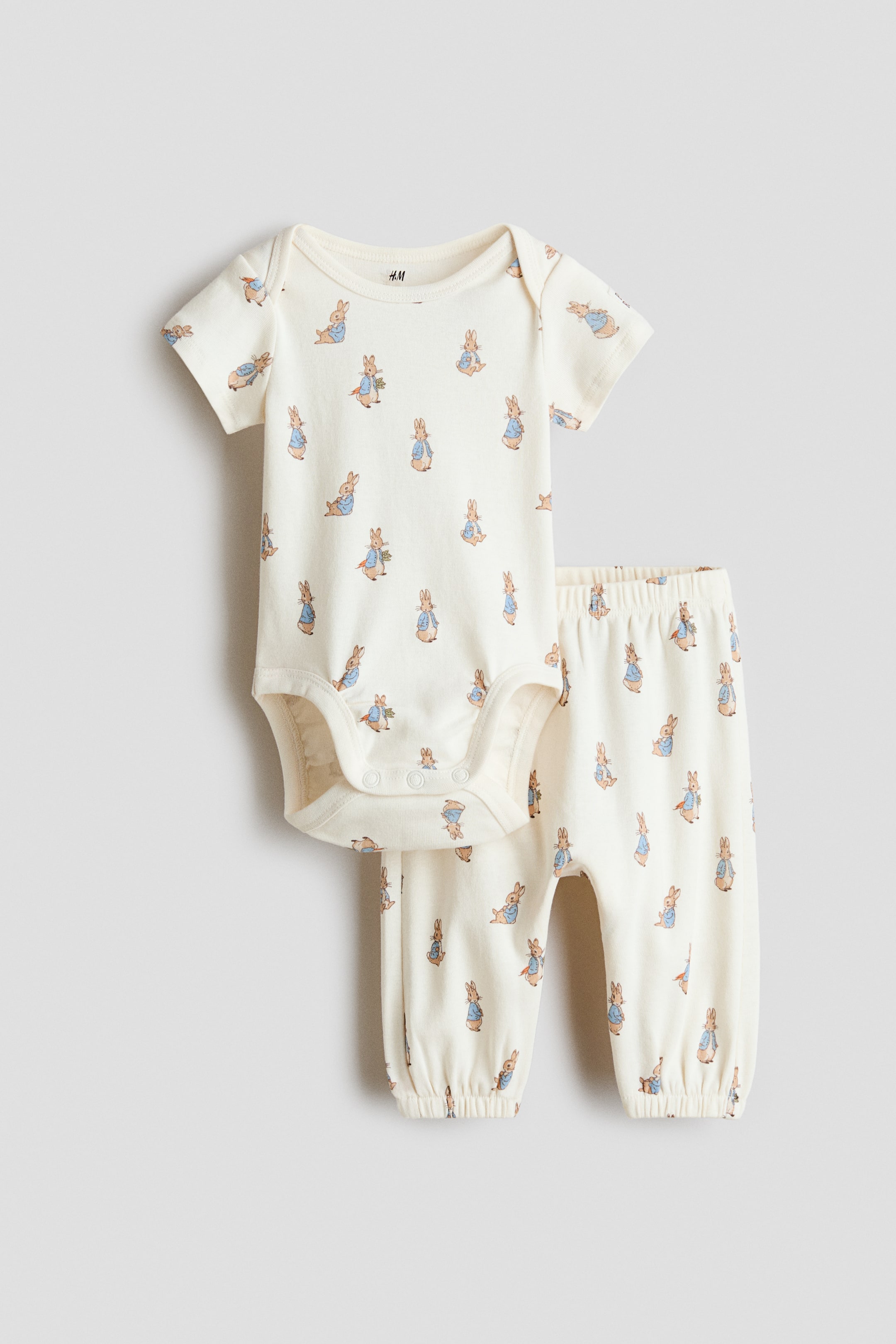 Voir une image plus grande: Un ensemble pour bébé crème présente un imprimé Peter Rabbit, face avant. L'ensemble comprend un body à manches courtes avec un col rond et des boutons-pression à l'entrejambe, ainsi qu'un pantalon long avec une taille élastiquée et des poignets élastiqués.