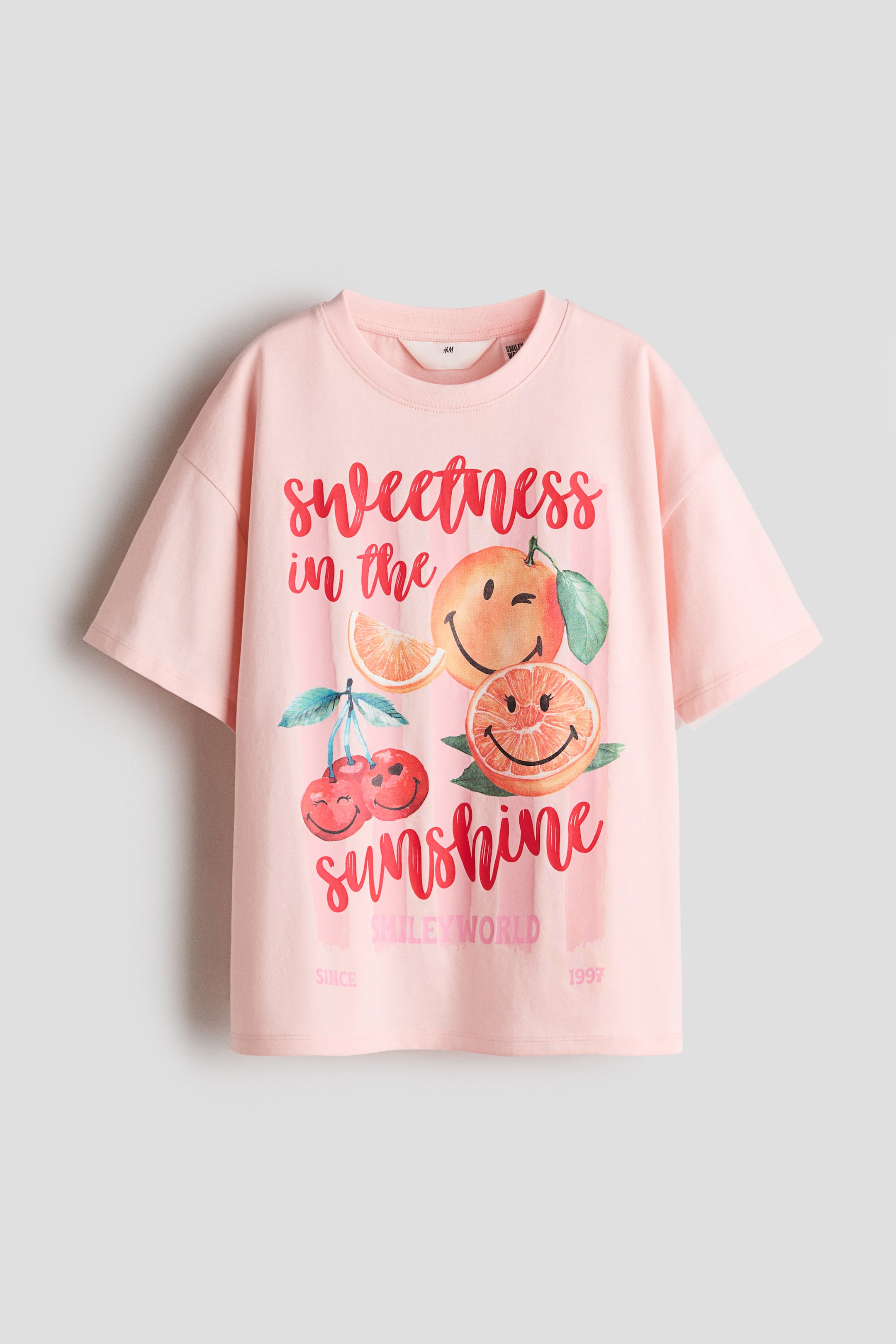 拡大画像を表示: オーバーサイズのピンクTシャツが前面を見せており、丸いリブトリムのネックラインとドロップショルダーが特徴です。プリントされたコットンジャージー素材で作られており、オレンジとチェリーのスマイリーフェイスのグラフィックと、赤いスクリプトフォントで「sweetness in the sunshine」というテキストが描かれています。