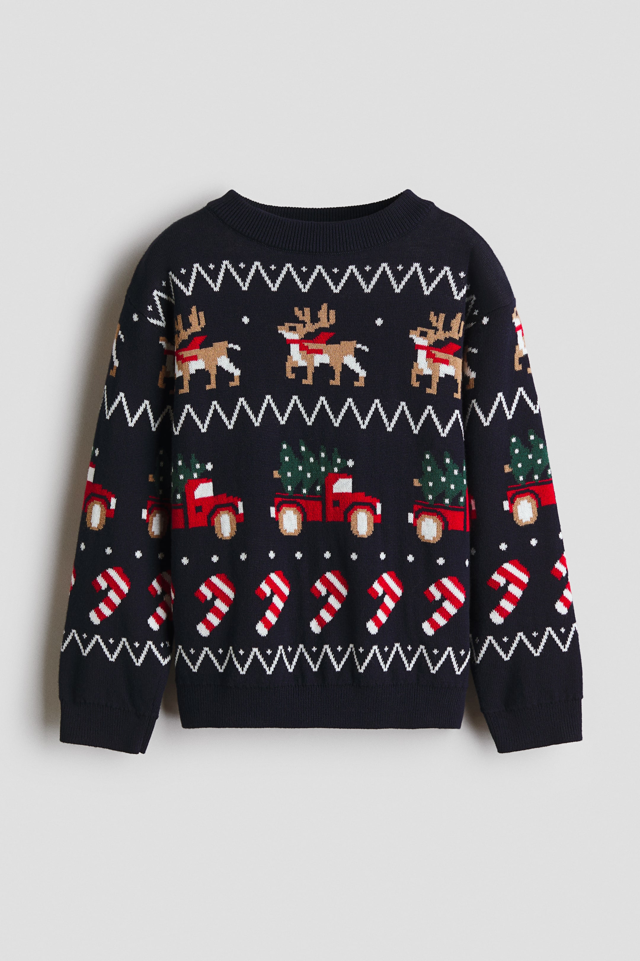 View larger image: A navy blue jacquard knit jumper is shown from the front, featuring horizontal bands of white zigzags, pixelated brown reindeer with red scarves, red pickup trucks carrying green Christmas trees, and red and white candy canes. It is designed with a round, rib-trimmed neckline, dropped shoulders, long sleeves, and ribbing at the cuffs and hem.