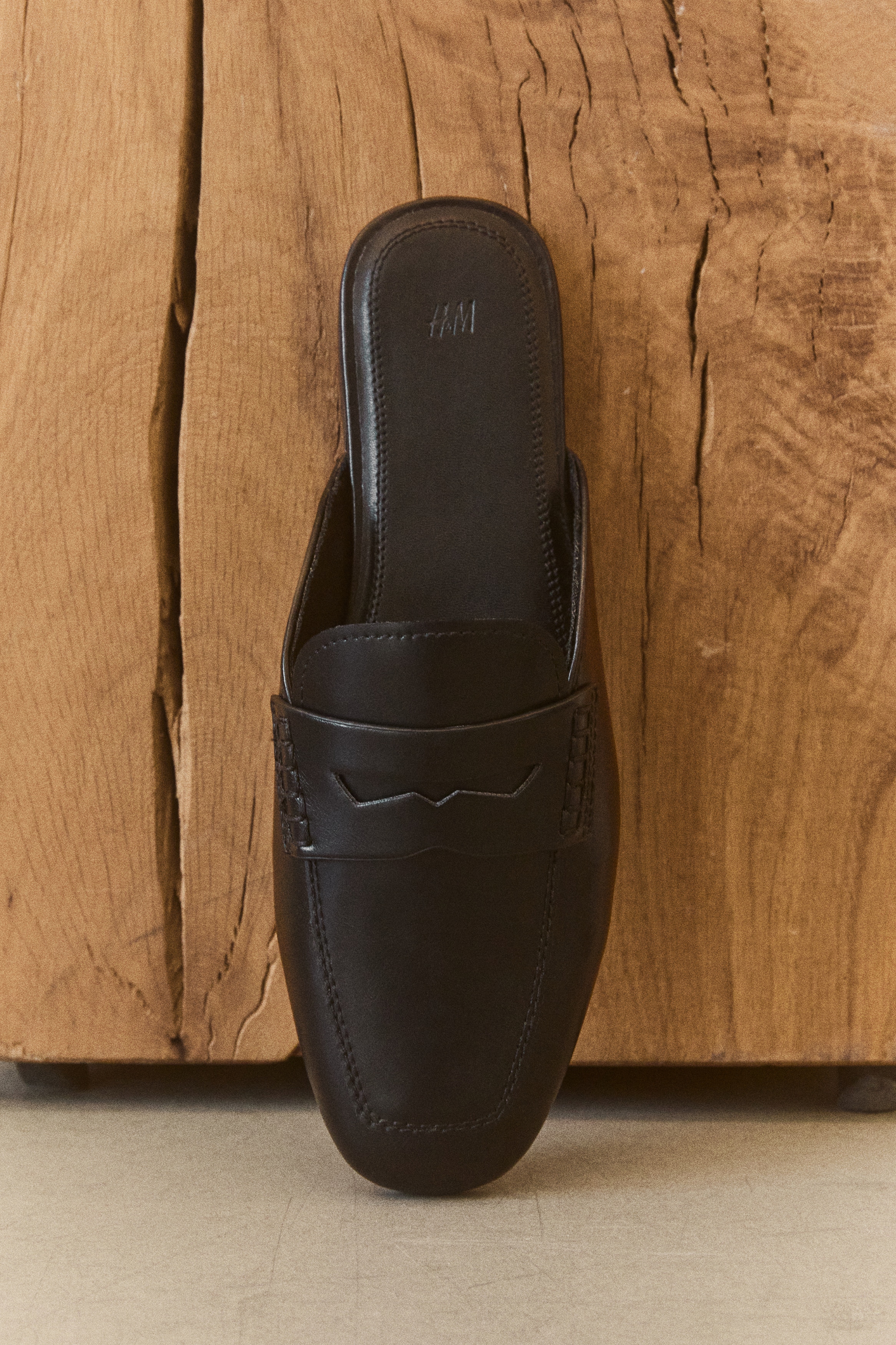 Two black loafer mules are facing forward, each featuring a smooth finish, a square-ish toe, and a classic loafer design. A saddle strap with a decorative cut-out crosses the instep, and visible stitching details the toe box. The H&M logo is embossed on the black insole of each shoe.