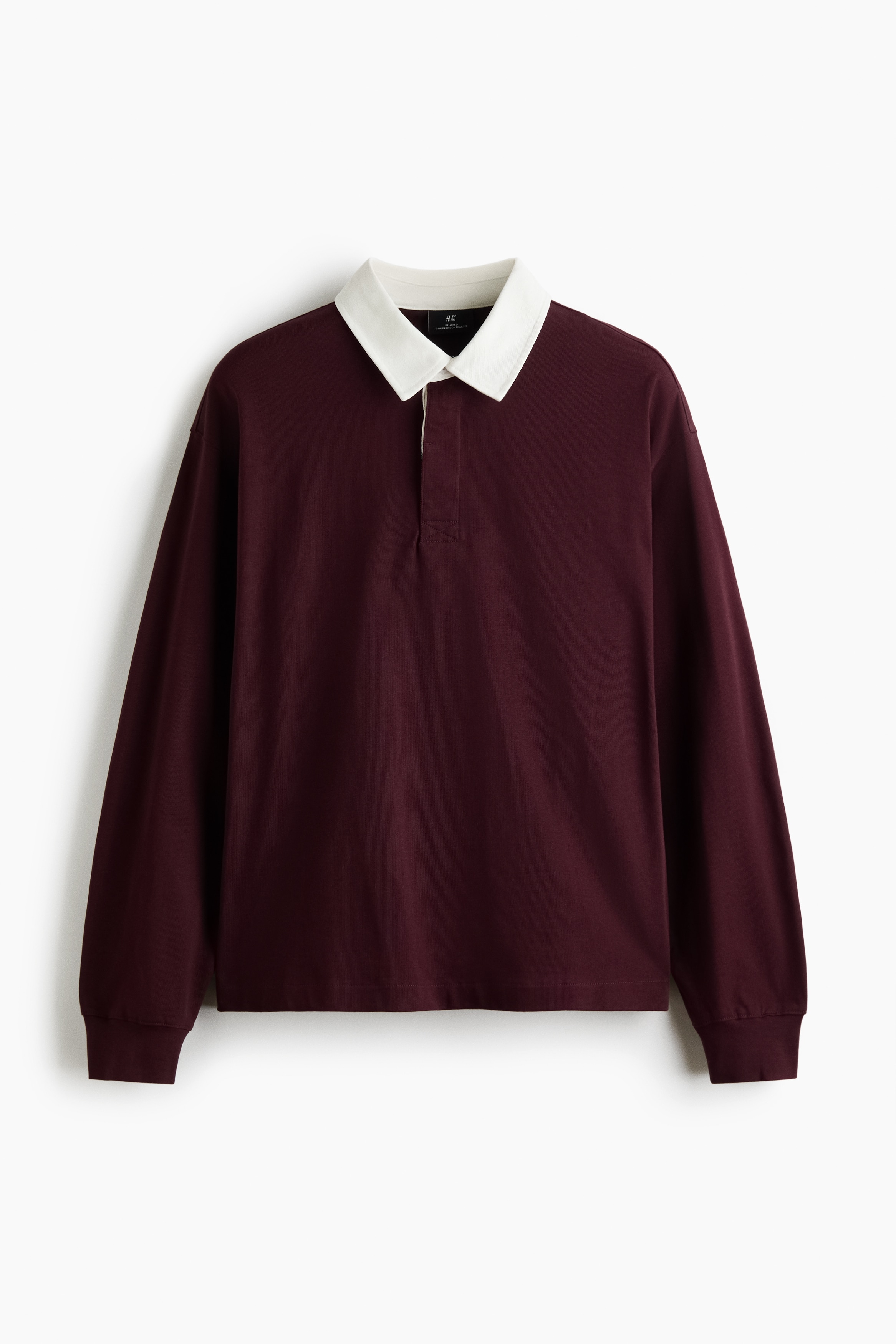 A maroon rugby shirt with a contrasting white collar and ribbed cuffs is being worn by the model over a white crew-neck T-shirt. The relaxed-fit shirt is paired with dark jeans, with the model placing one hand in a front pocket.