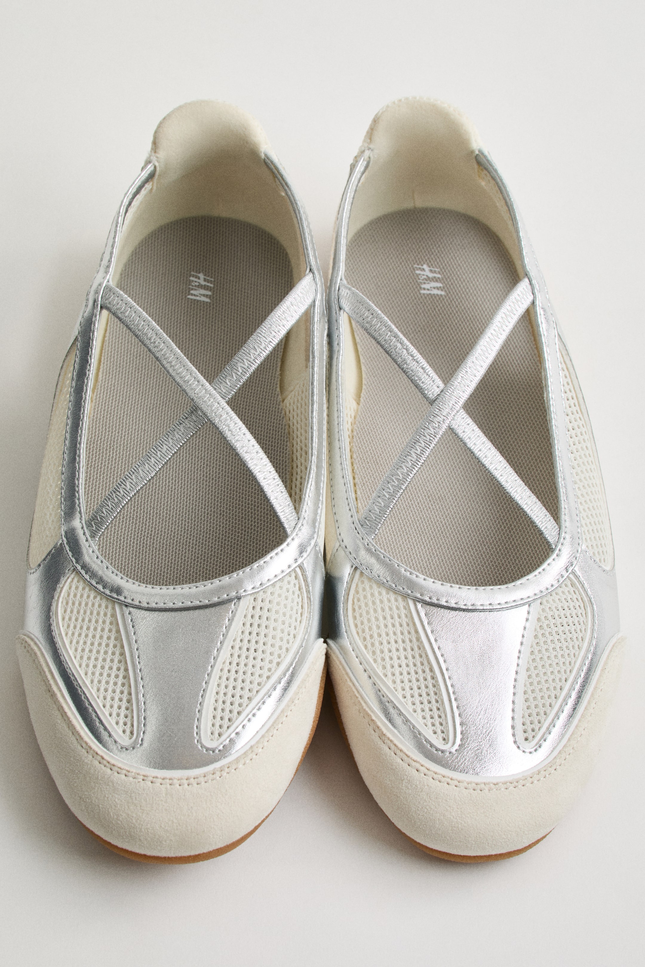 View larger image: A pair of silver and white slip-on shoes is facing forward, featuring white mesh panels and metallic silver accents throughout. Criss-crossing elastic straps cover the instep, and light beige suede-like material forms the toe caps and parts of the sides.