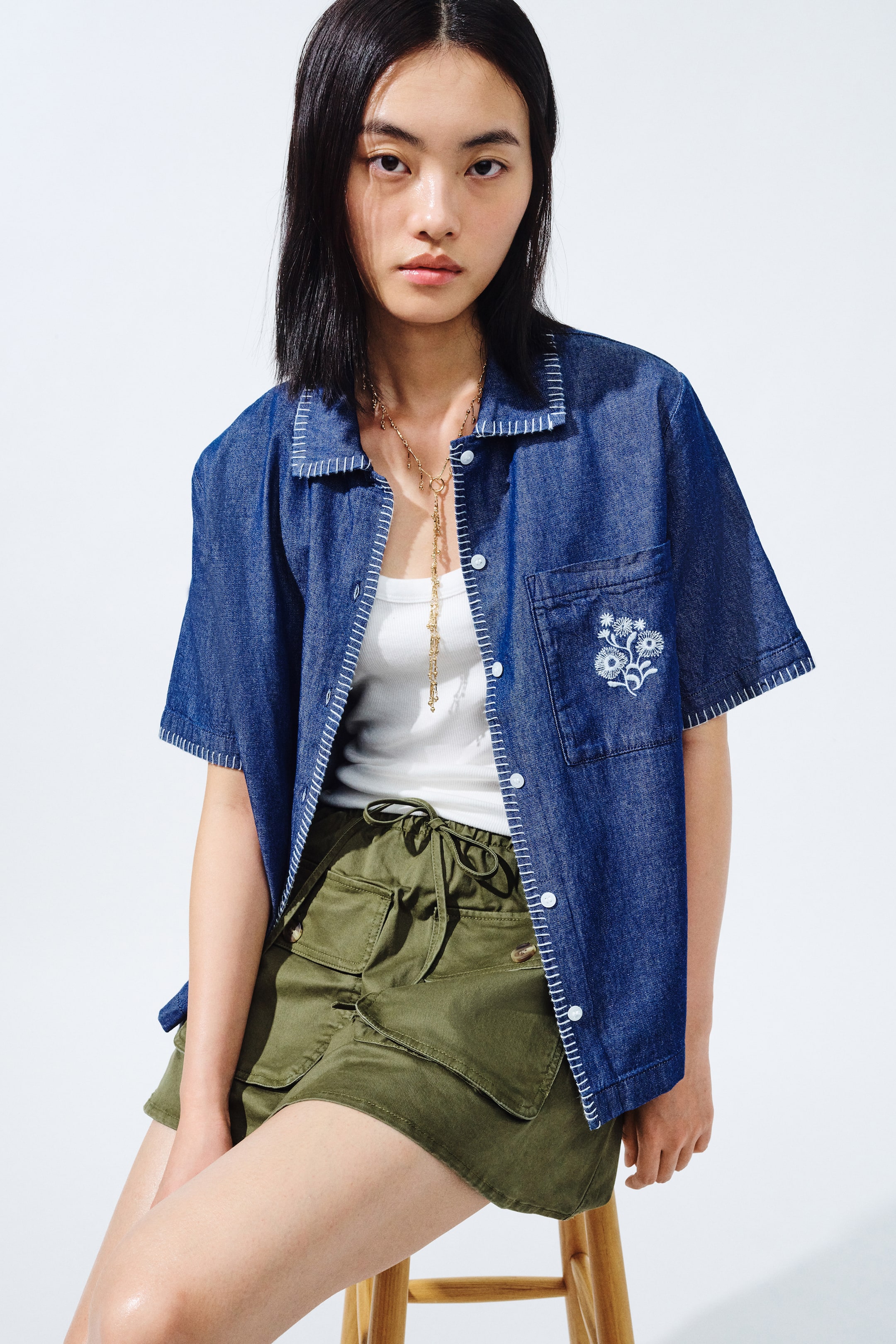 View larger image: A blue denim short-sleeved shirt showcases white contrast stitching along its seams, a pointed collar, white buttons, and a white floral embroidery on its left chest pocket. It is styled open over a white tank top and paired with olive green utility shorts, featuring an elasticated drawstring waistband and large flap pockets on the front.