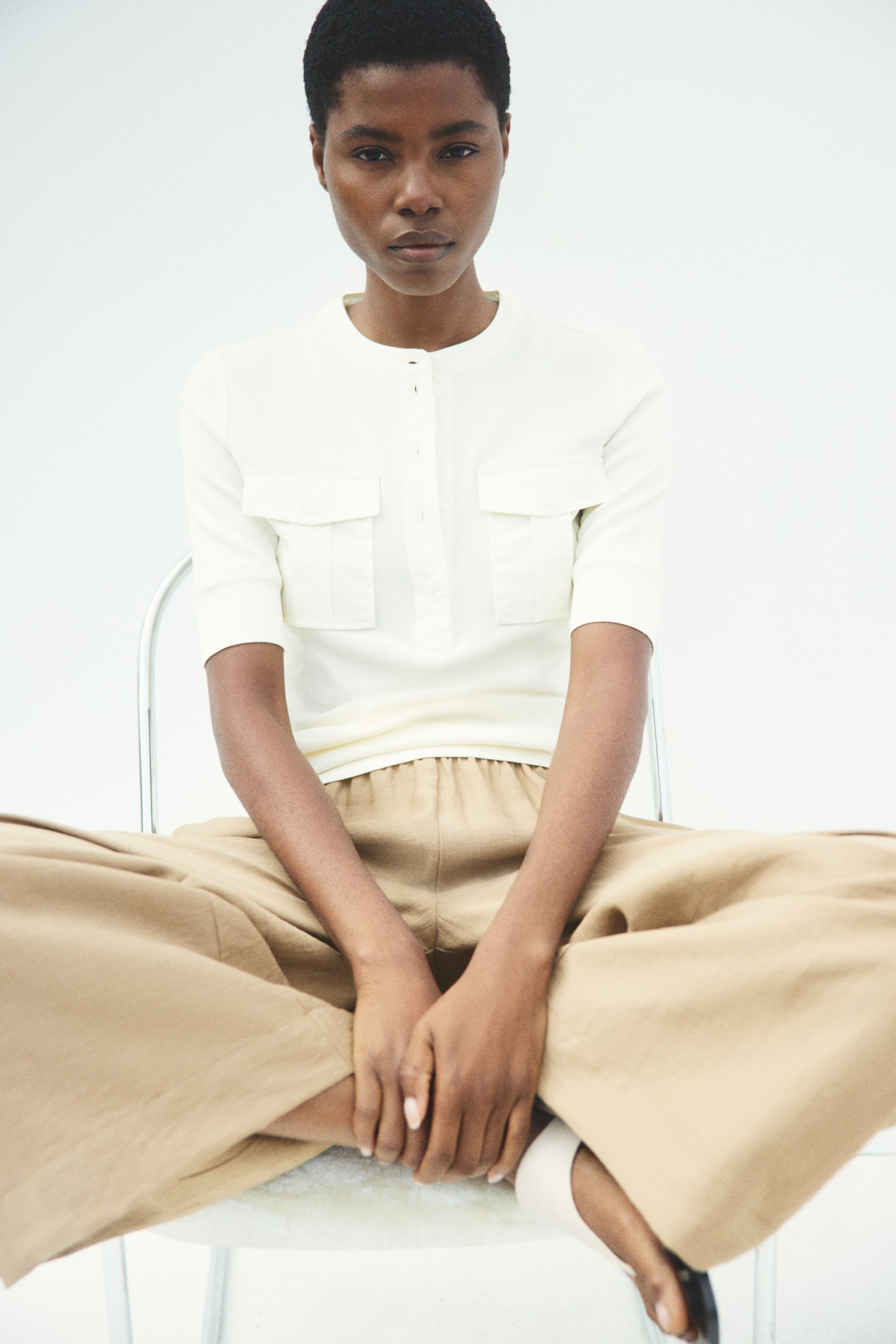 View larger image: Loose-fit, ankle-length tan trousers with wide legs are being worn by the model, featuring a high, elasticated waist. The model is sitting with their legs crossed, showcasing the relaxed drape of the fabric.