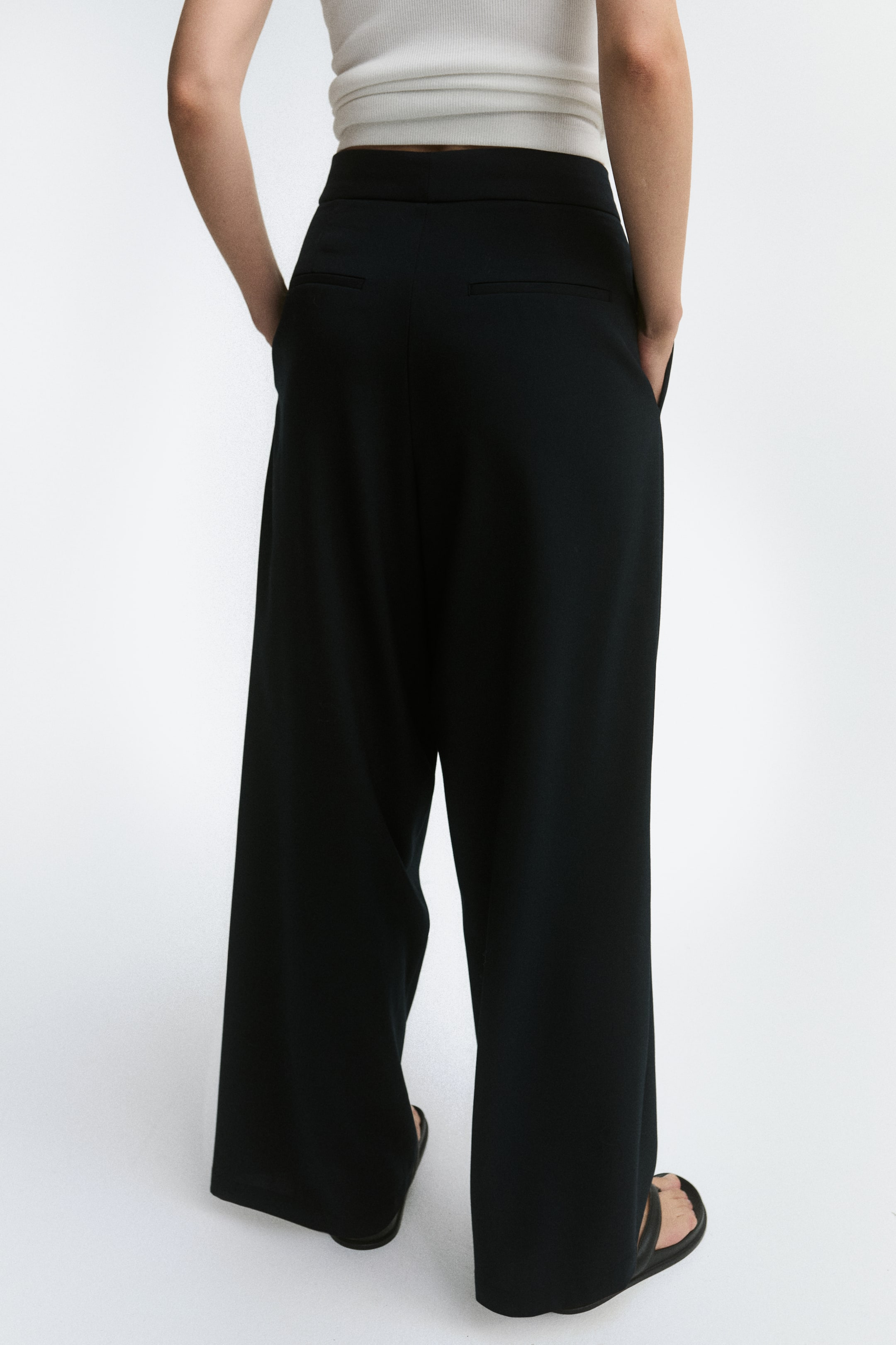View larger image: Loose-fit black pants in a woven fabric are shown from the back, highlighting a high waist, a structured waistband, and two jetted back pockets. The pants drape into a relaxed barrel-leg silhouette and are paired with a tucked-in white top.