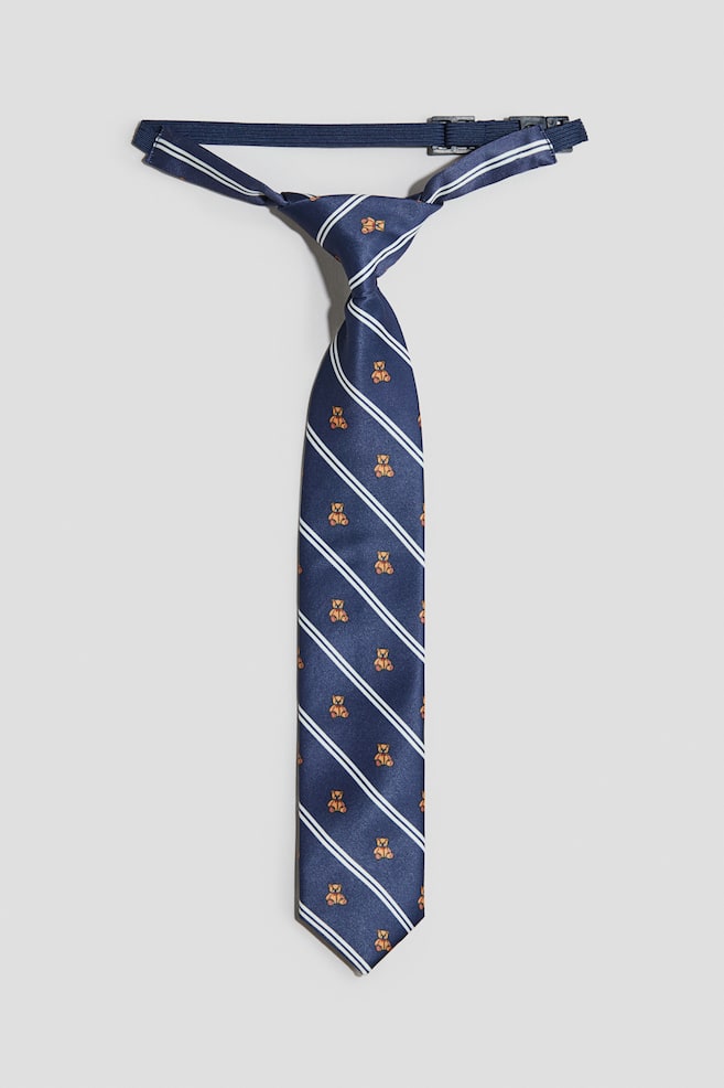 Se muestra de frente una corbata de satÃ©n azul marino ya anudada, con un diseÃ±o de dobles franjas diagonales blancas y pequeÃ±os ositos de peluche naranjas. Cuenta con una banda elÃ¡stica oscura ajustable y un broche de plÃ¡stico en la parte trasera. - 1