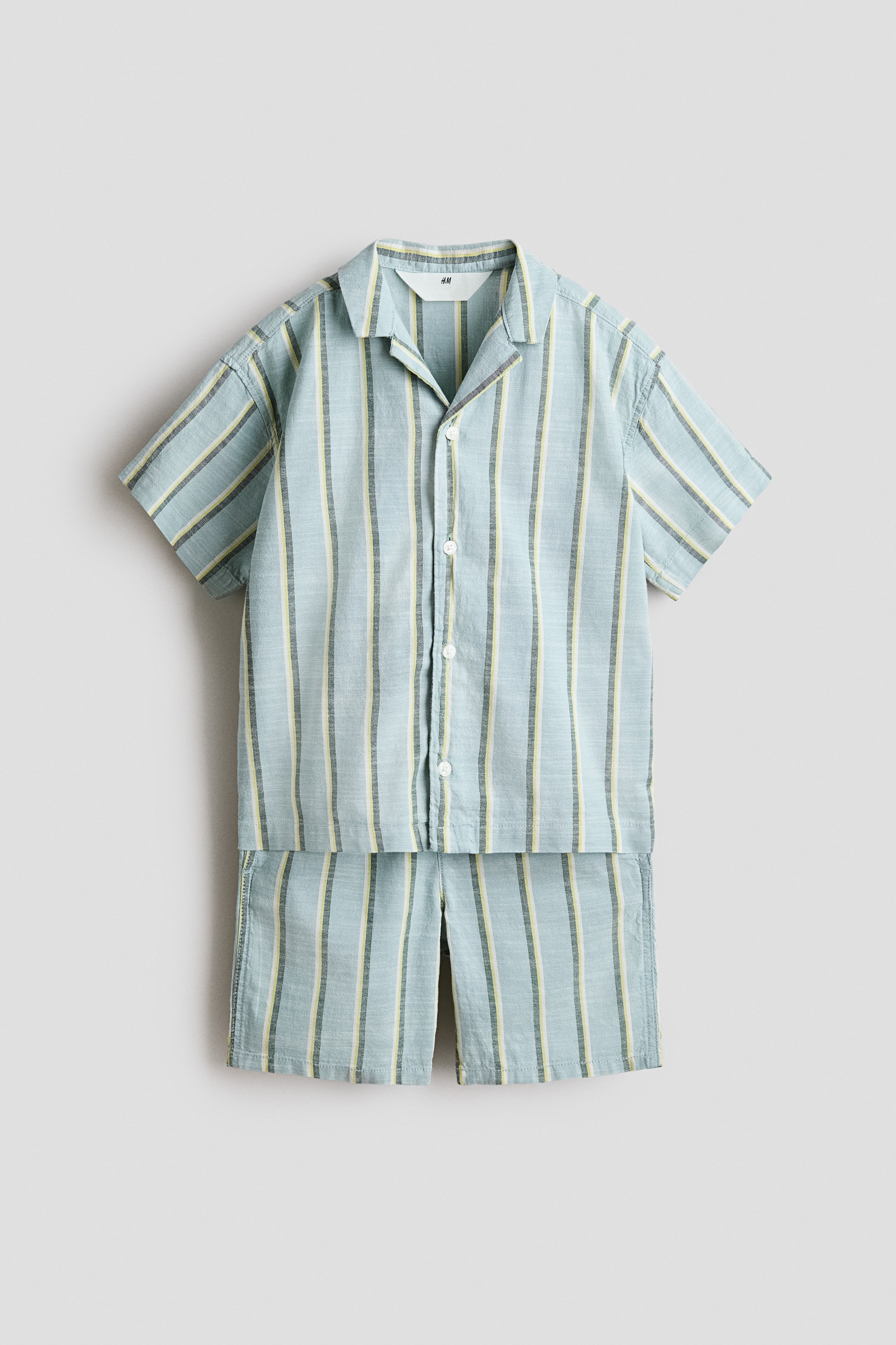 View larger image: A front-facing set features a resort shirt and a pair of shorts, both made from a light-blue, airy cotton weave with vertical stripes in yellow and dark grey. The short-sleeved shirt features a resort collar, gently dropped shoulders and buttons down the front, while the shorts are designed with an elasticated drawstring waist and discreet side pockets.