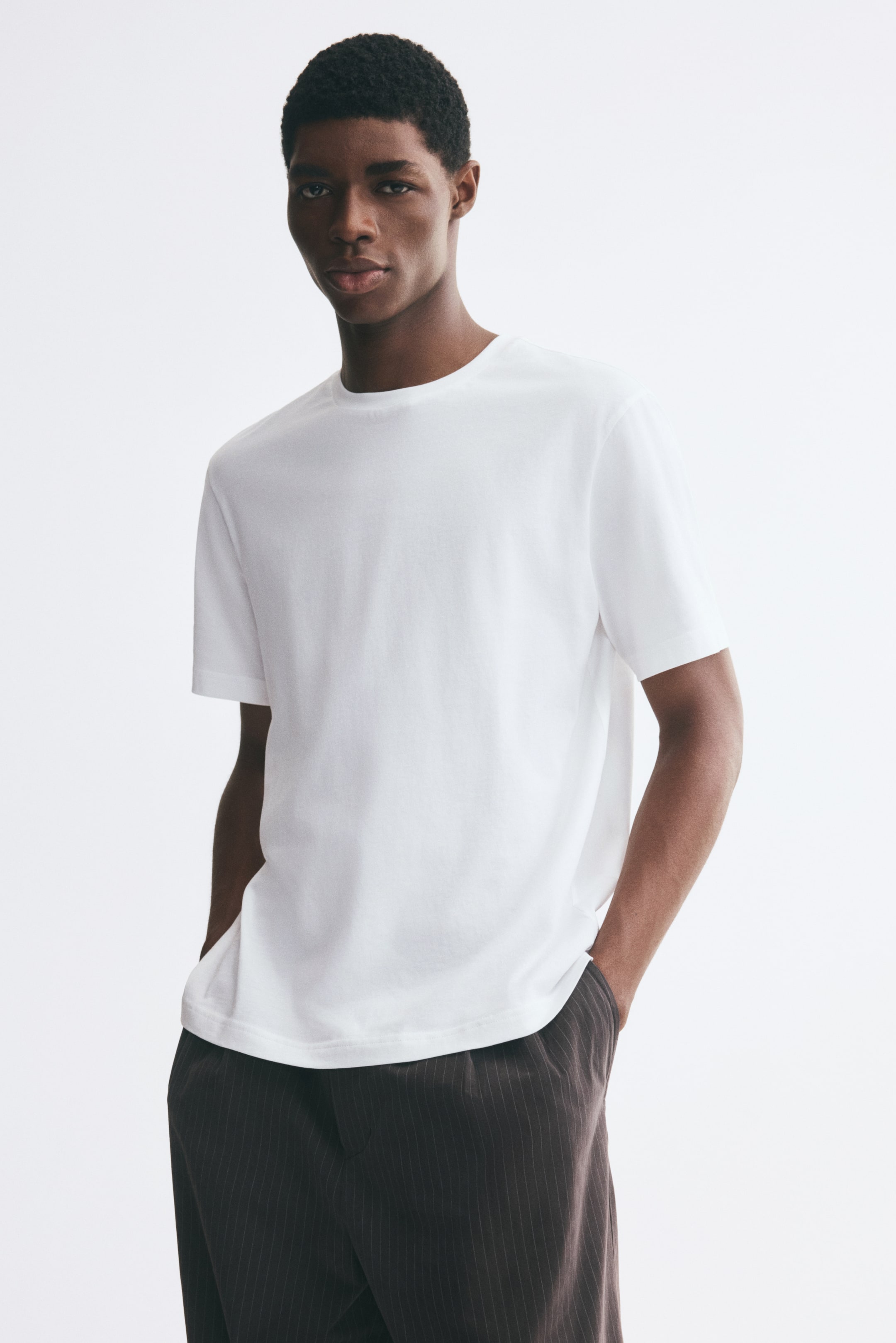 Größeres Bild anzeigen: Das Model trägt ein weißes T-Shirt mit Rundhalsausschnitt und kurzen Ärmeln. Es steht mit den Händen in den Taschen einer dunklen, fein gestreiften Hose und präsentiert so die lockere Passform und den geraden Saum des T-Shirts.