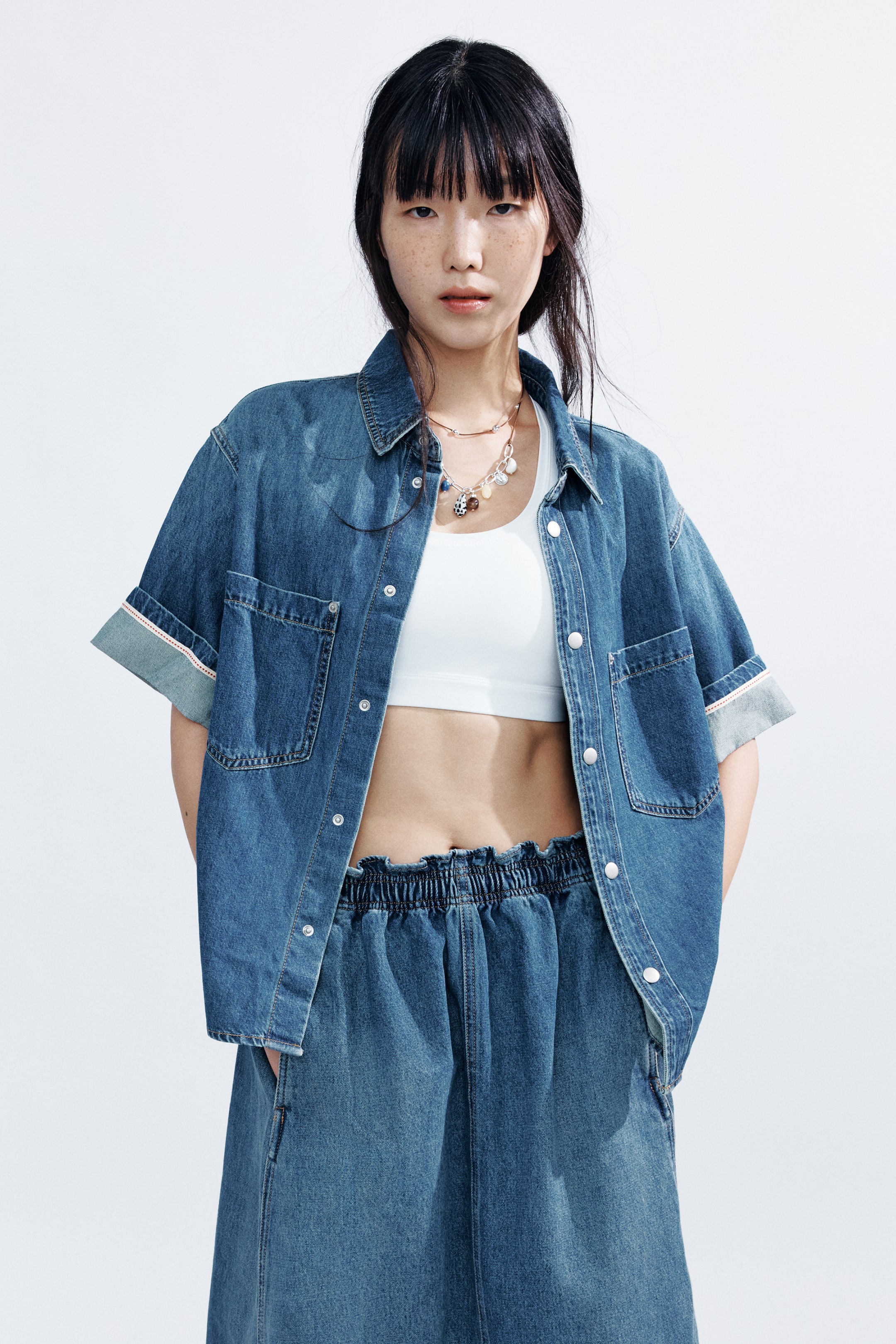 View larger image: A loose-fit cotton denim shirt with a collar, white press-studs, patch chest pockets, dropped shoulders, and turn-up cuffs is being worn open. The model is styling the shirt over a white bra top.
