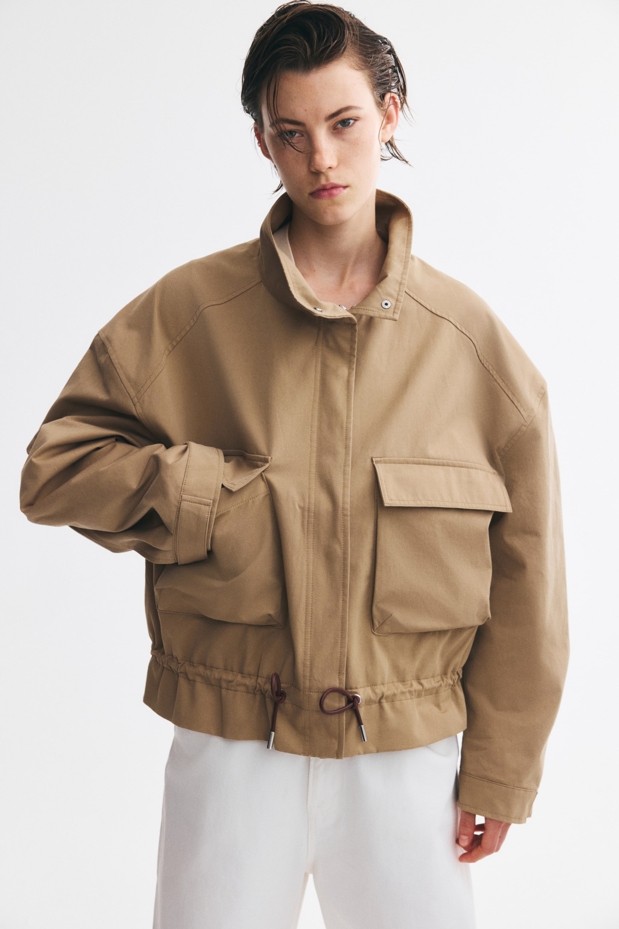View larger image: An oversized beige twill jacket is shown from the front, featuring a high funnel collar, dropped shoulders, and a concealed front fastening. The jacket features large bellows pockets with flaps and a gathered hem with a dark brown drawstring.