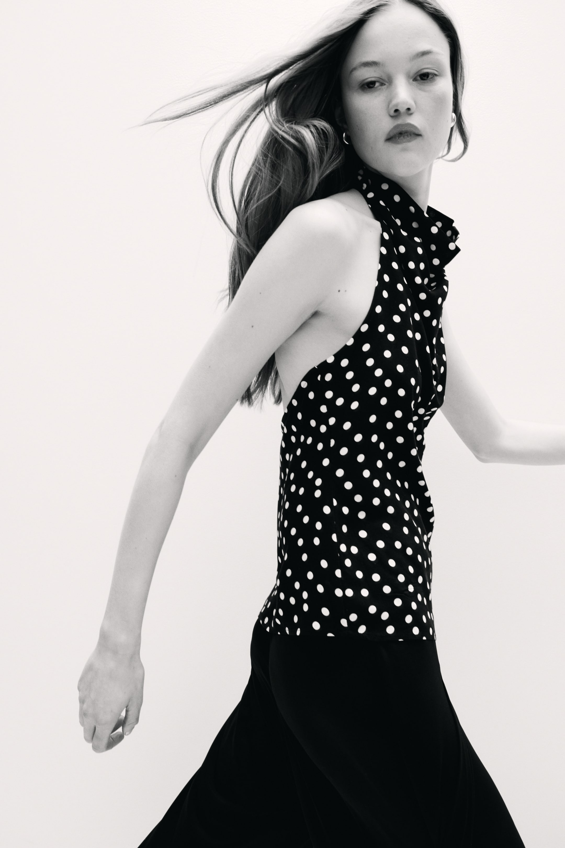 View larger image: A black halter neck top, adorned with white polka dots and a high, ruffled neckline, is displayed from a side-front perspective, highlighting its open side design. Black trousers with a flowing silhouette are worn underneath.