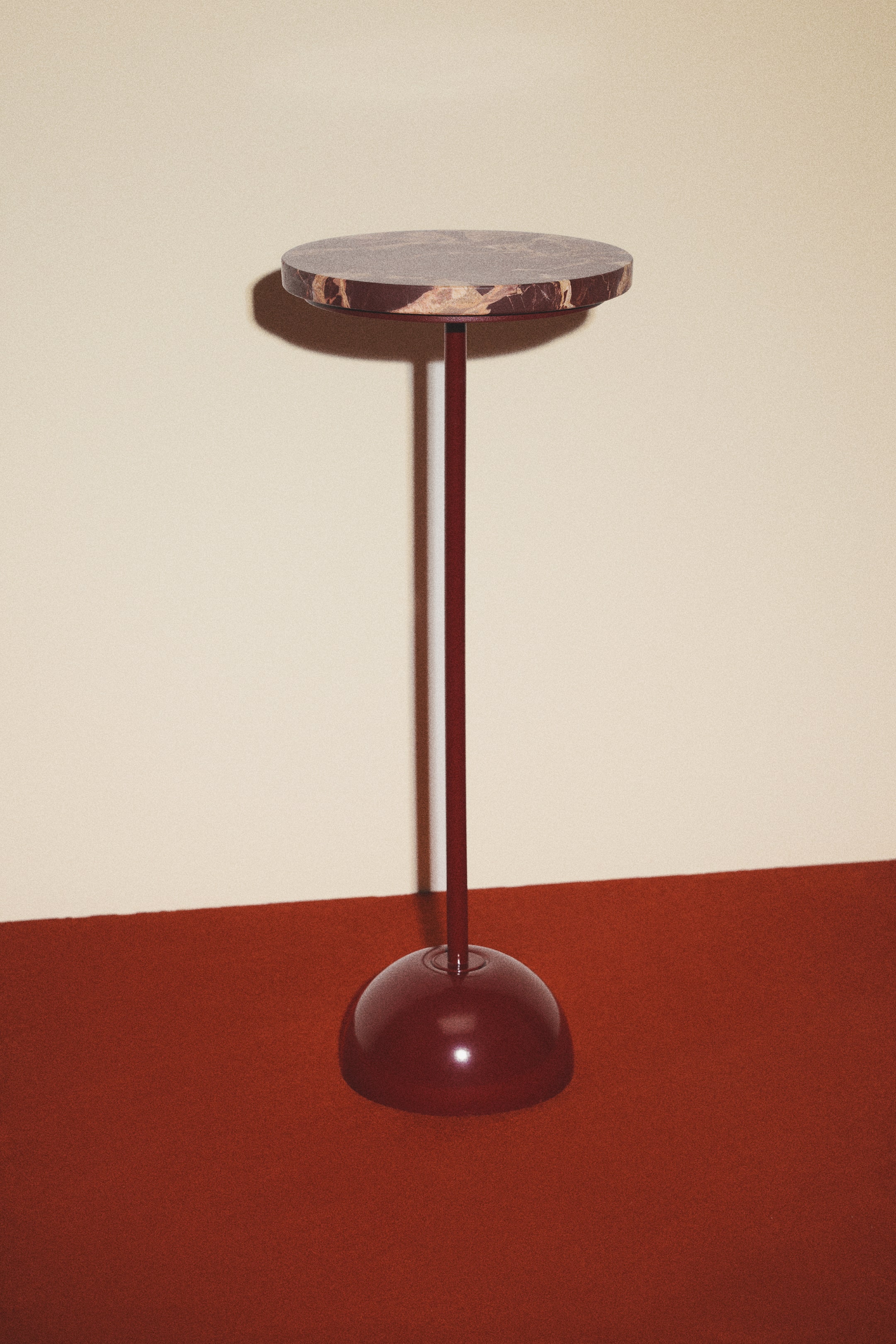 View larger image: A pedestal table is standing, featuring a round, brown marble slab top with lighter veining. It is supported by a slender, dark red metal pole connecting to a matching dome-shaped metal base.