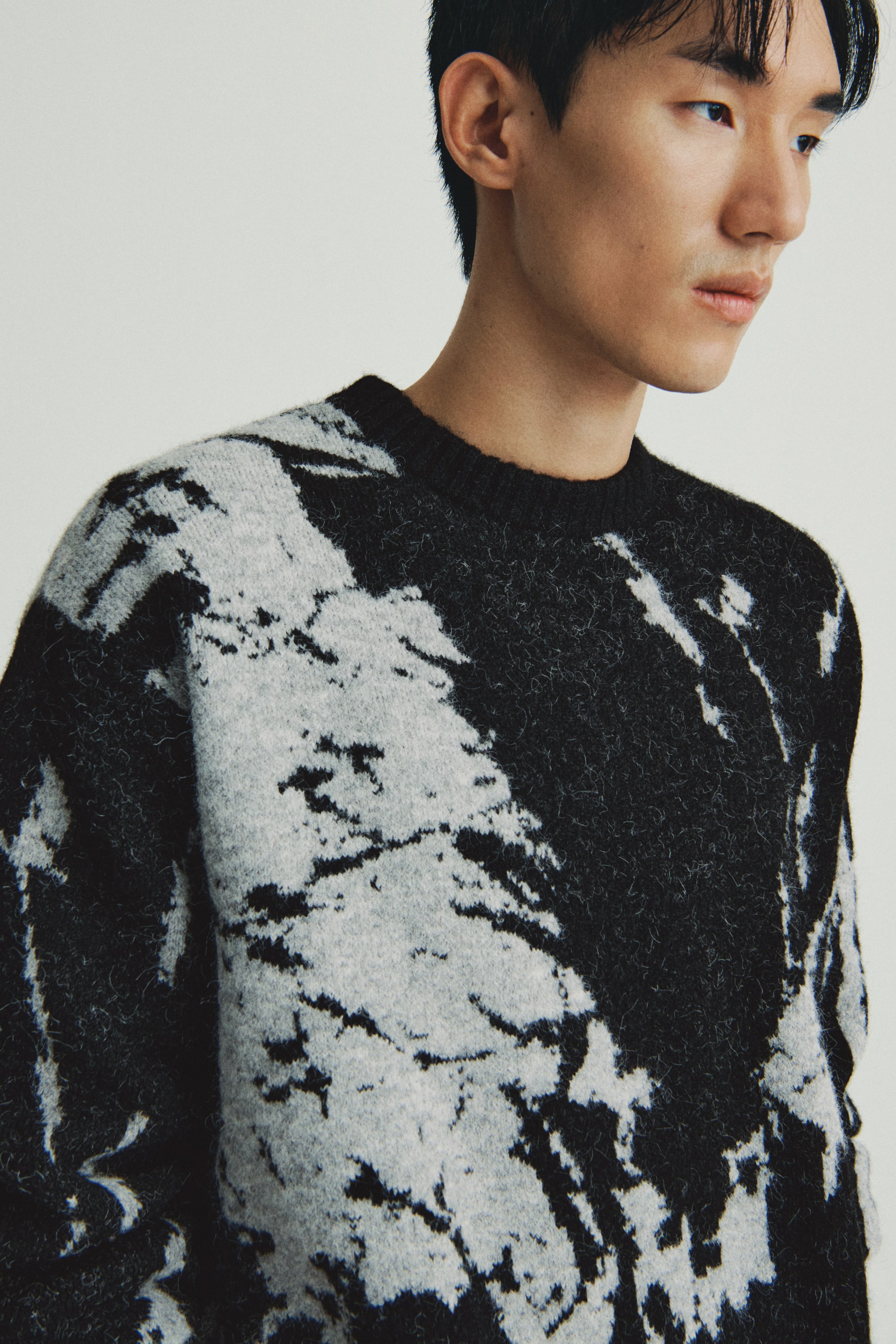 이미지 확대 보기: A crew neck sweater is being shown from the front and right shoulder, featuring a black and light gray abstract, mottled pattern. 부드럽고 브러시드된 텍스처를 가진 것으로 보입니다.