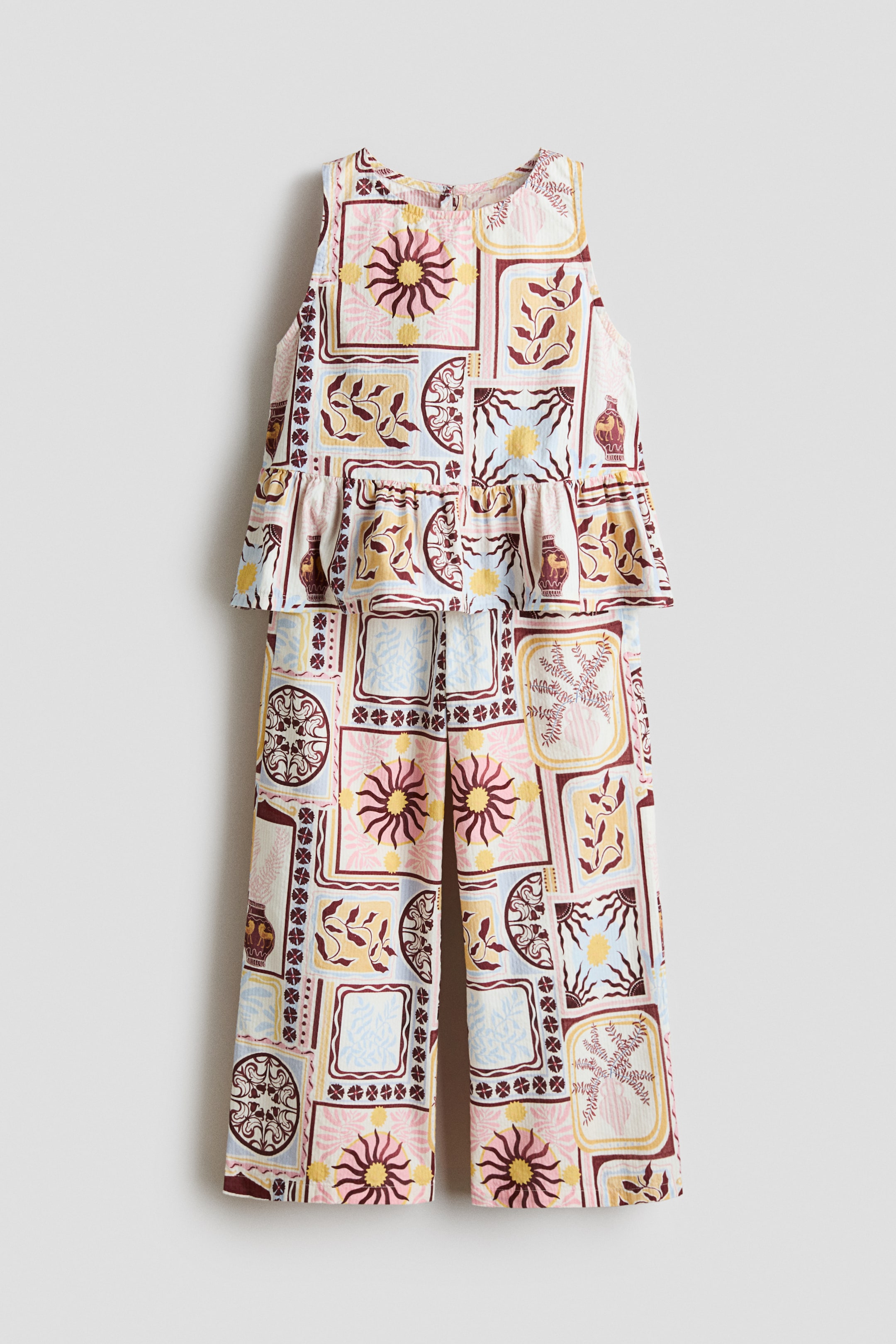 View larger image: A sleeveless children's jumpsuit shown from the front, featuring a round neckline, a ruffled peplum waist and wide-leg trousers, all displaying an all-over mosaic pattern of suns, vases, leaves and geometric shapes in pink, yellow, blue and maroon.