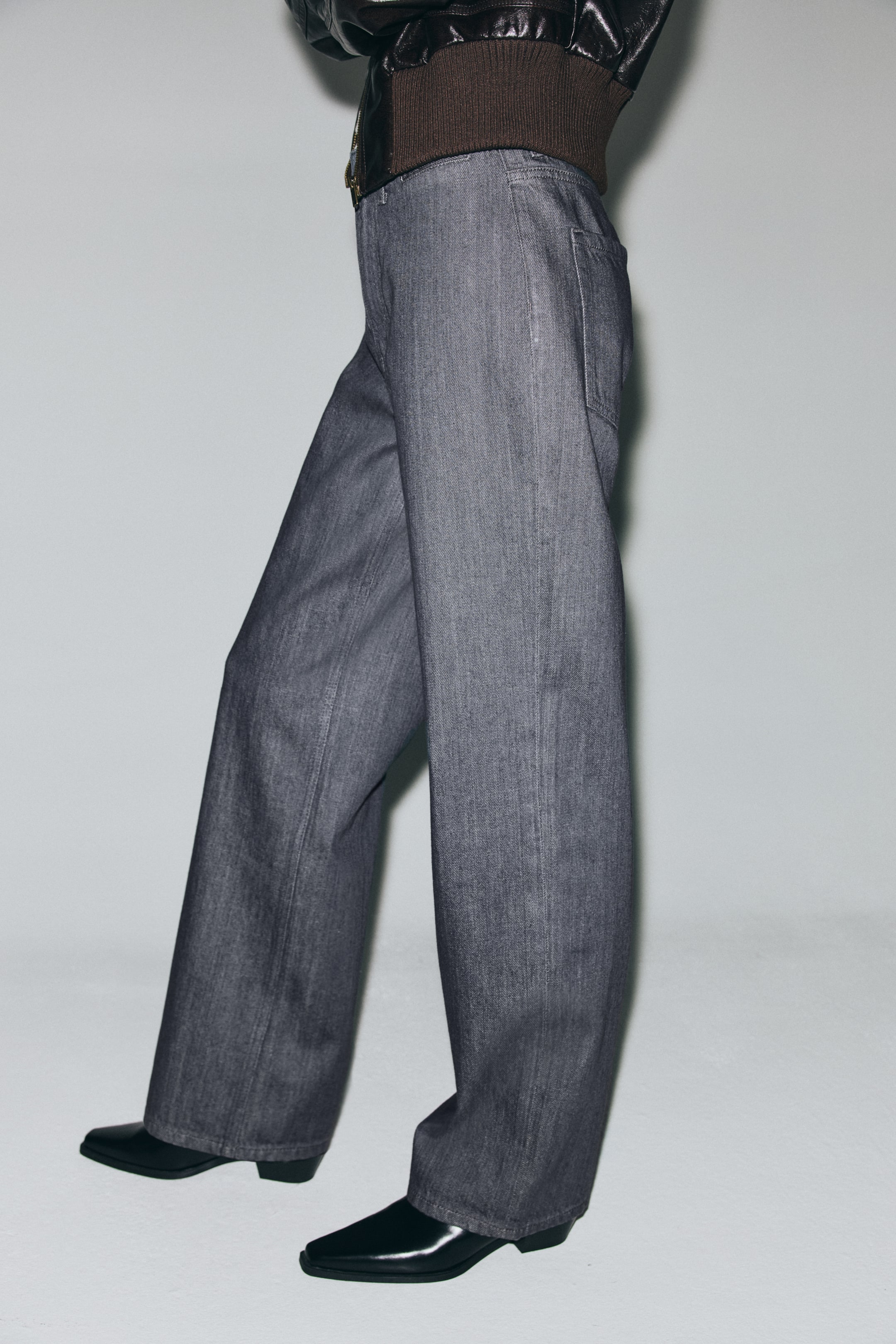 Agrandir l'image: Des jeans gris foncé à jambes larges sont portés, avec une taille haute partiellement recouverte par une ceinture côtelée marron foncé, une poche arrière visible et des coutures verticales proéminentes courant sur toute la longueur des jambes. Les jeans s'accumulent légèrement sur des bottes noires à bout pointu.