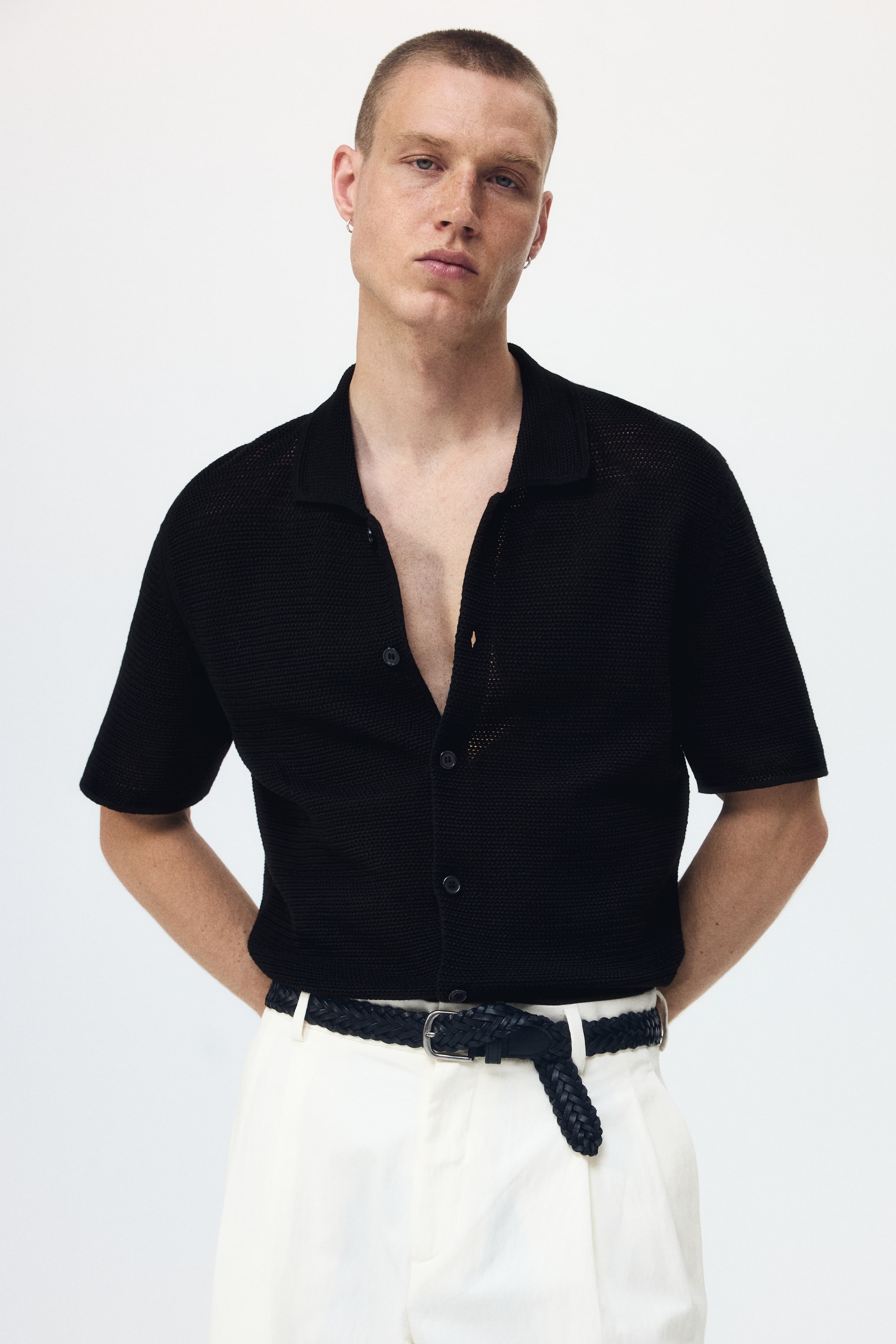 Ver imagen más grande: La camiseta polo de punto de manga corta en color negro se presenta en una vista frontal, con un patrón de punto texturizado y un frente abotonado con botones oscuros. La camiseta está parcialmente desabrochada y metida dentro del pantalón, revelando unos pantalones blancos y un cinturón trenzado negro.