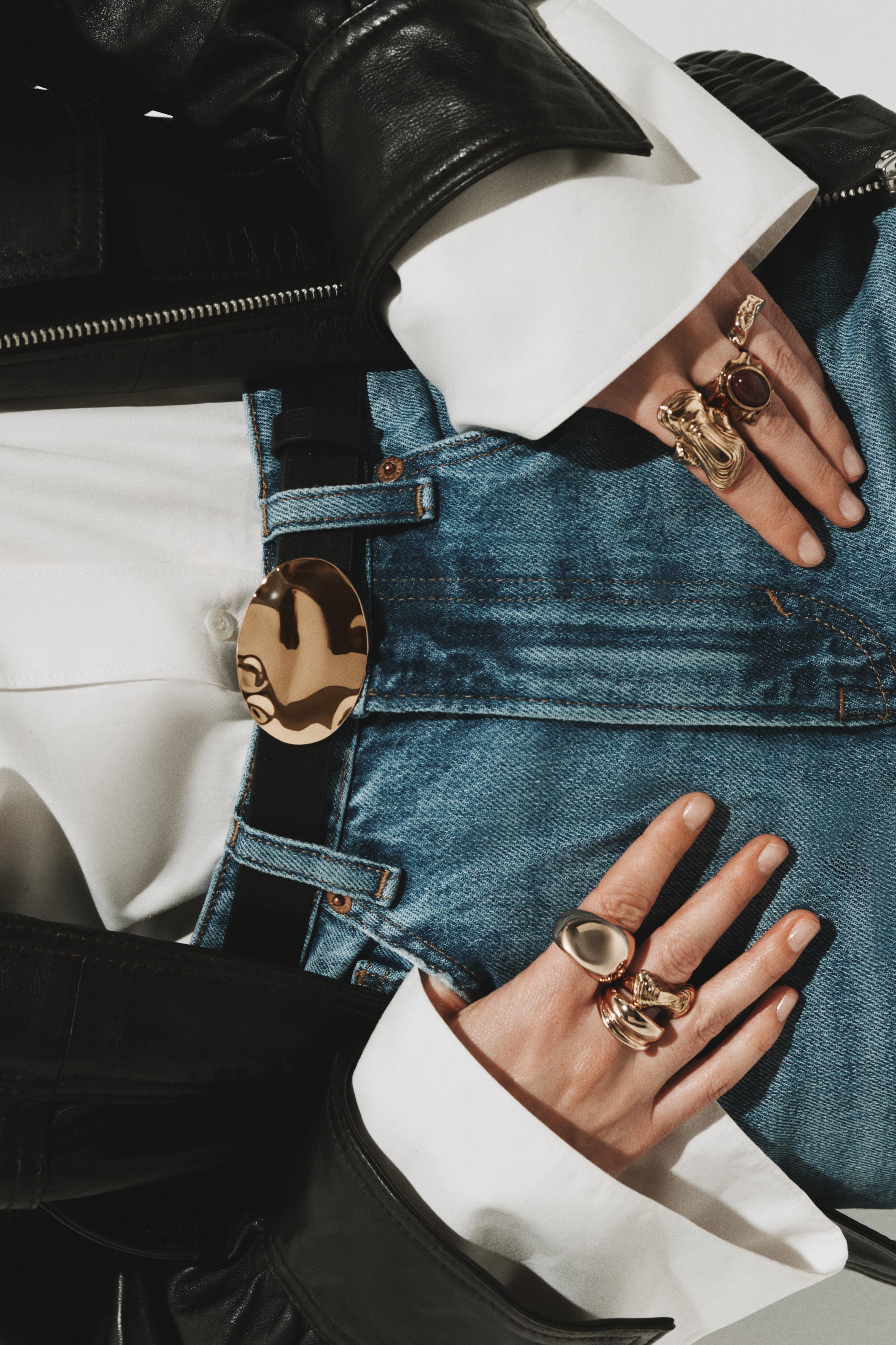 View larger image: A black leather jacket layered over a white button-down shirt with wide cuffs, tucked into blue denim jeans. A black belt with a large, irregular gold buckle cinches the jeans; hands rest on the waistband, displaying multiple gold rings in sculptural and stone-set designs.