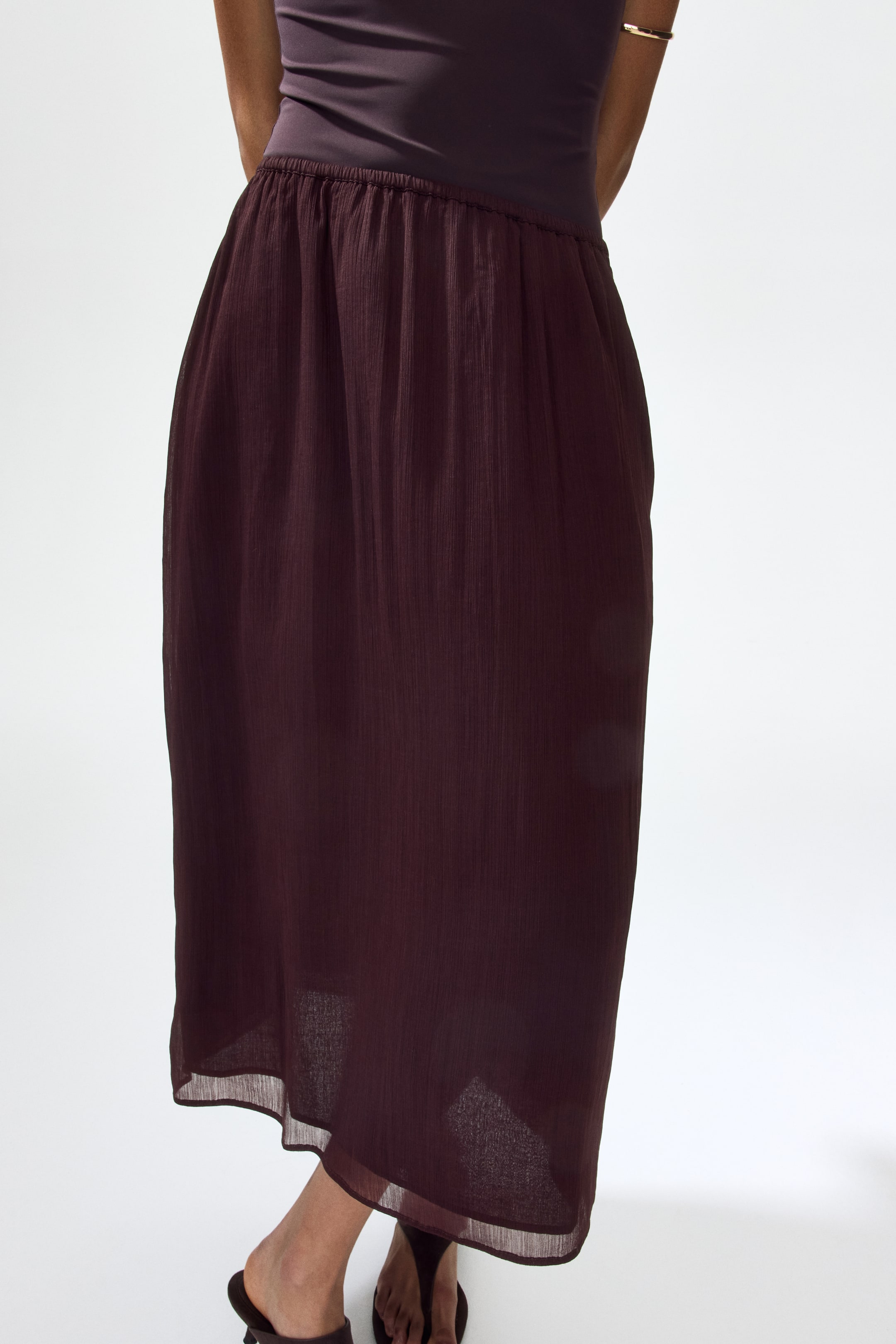 View larger image: "A deep maroon midi skirt is displayed from the back, showcasing a gathered elasticated waistline positioned beneath a snug top. Crafted from a crinkled, sheer fabric, it drapes gracefully down to mid-calf length."