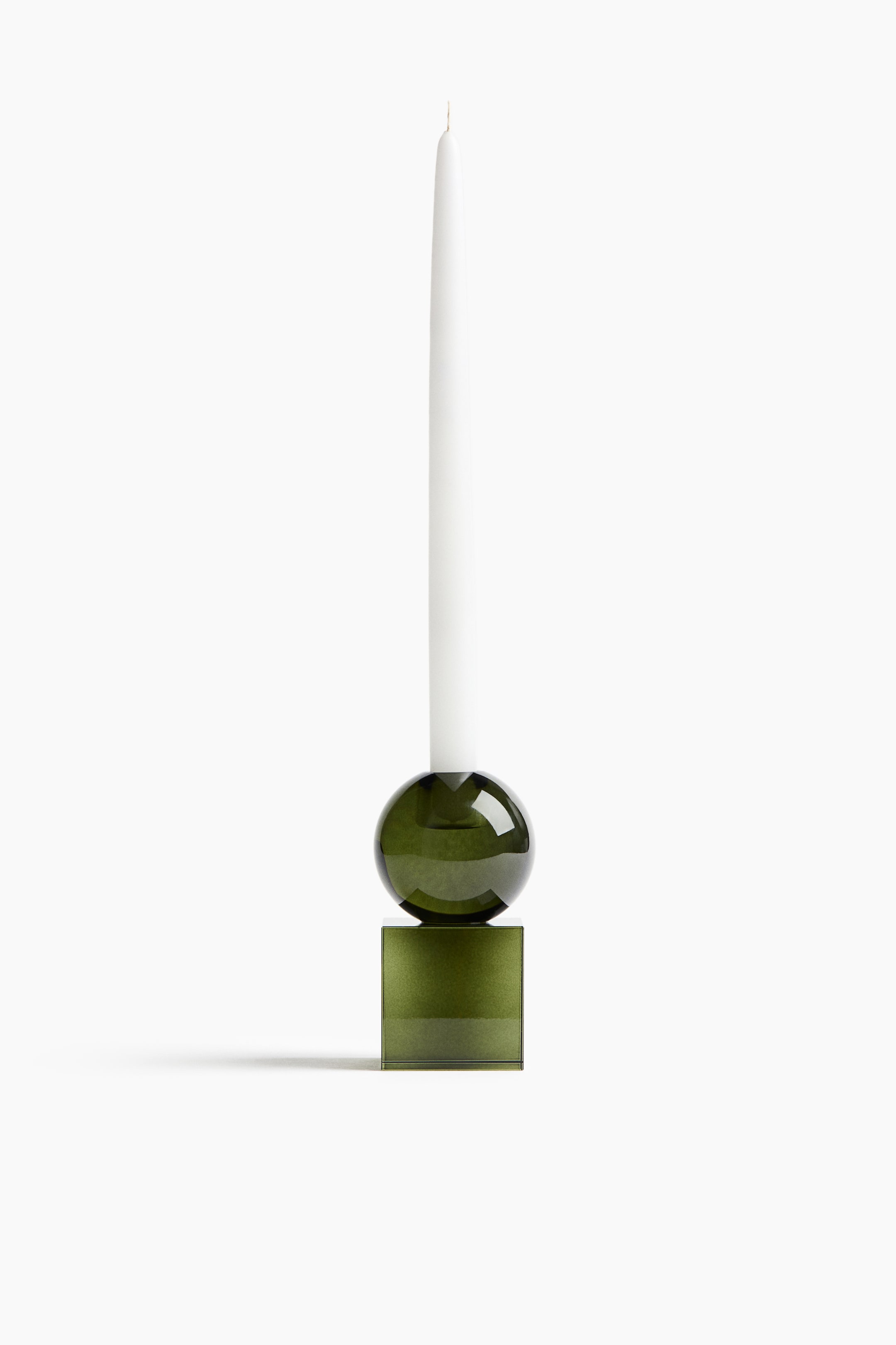 View larger image: A green glass candle holder, shown from a front angle, supports a tall white taper candle. The candle holder features a modern geometric design with a translucent olive-green cube base stacked beneath a matching glass sphere.