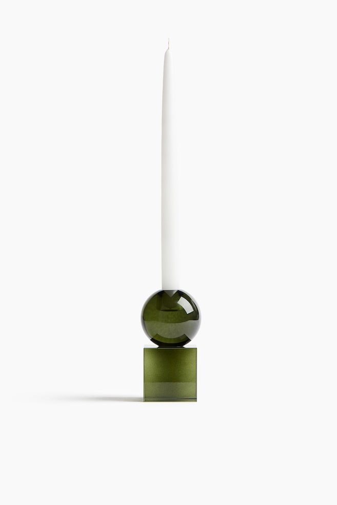 A green glass candle holder, shown from the front, supports a tall white taper candle. The candle holder features a modern geometric design, with a translucent olive-green cube base topped by a matching glass sphere. - 1