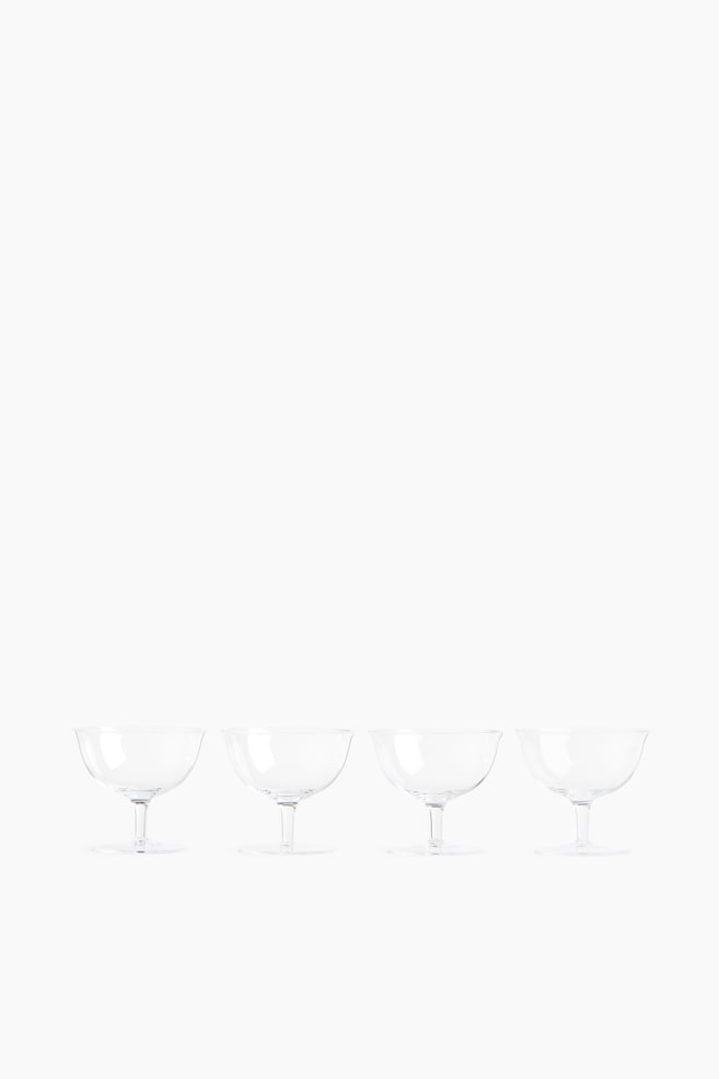 Quatro taÃ§as de quatro pÃ©s em vidro transparente e delicado estÃ£o dispostas numa fila horizontal, viradas para a frente. Cada taÃ§a apresenta uma forma suavemente arredondada, com rebordo ligeiramente aberto, apoiada por um pÃ© curto e robusto e uma base circular plana. - 1