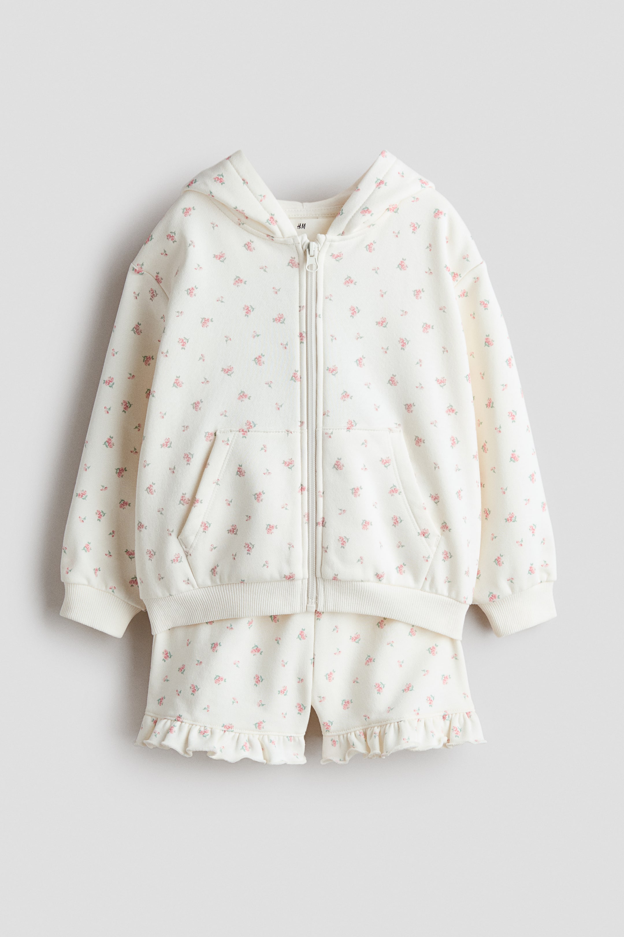 Visualizza immagine più grande: Questo set presenta una felpa con cappuccio e zip abbinata a shorts, mostrati in primo piano, entrambi in tessuto felpato bianco sporco con una delicata stampa floreale rosa e verde. La felpa con cappuccio è dotata di cappuccio foderato in jersey, zip frontale, spalle cadenti e tasche anteriori, con bordi a coste ai polsini e all'orlo, mentre gli shorts sono progettati con vita elasticizzata e bordo con volant agli orli.