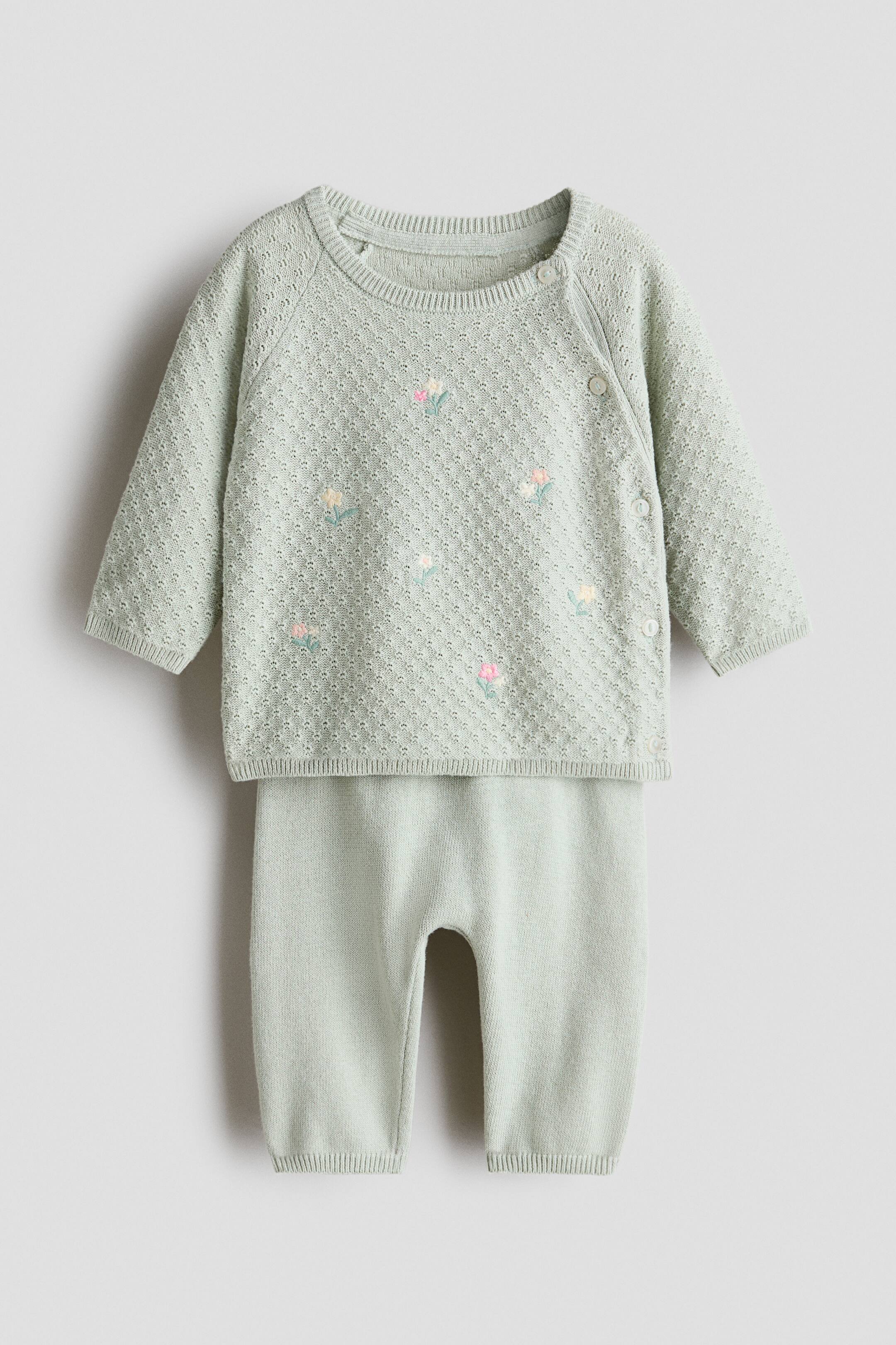 Näytä suurempi kuva: Vaaleanvihreä kaksiosainen neulottu setti vauvoille näkyy EDESTÄ, sisältäen puseron ja yhteensopivat housut. Puserossa on jacquard-neulos, hajallaan olevia kukkakirjailuja, pyöreä pääntie, pitkät raglanhihat ja sivunapin kiinnitys, kaikki viimeistelty resoreilla hihansuissa ja helmassa; housuissa on esillä sileä joustava vyötärö, leveä lantio ja resoritetut hihansuut.
