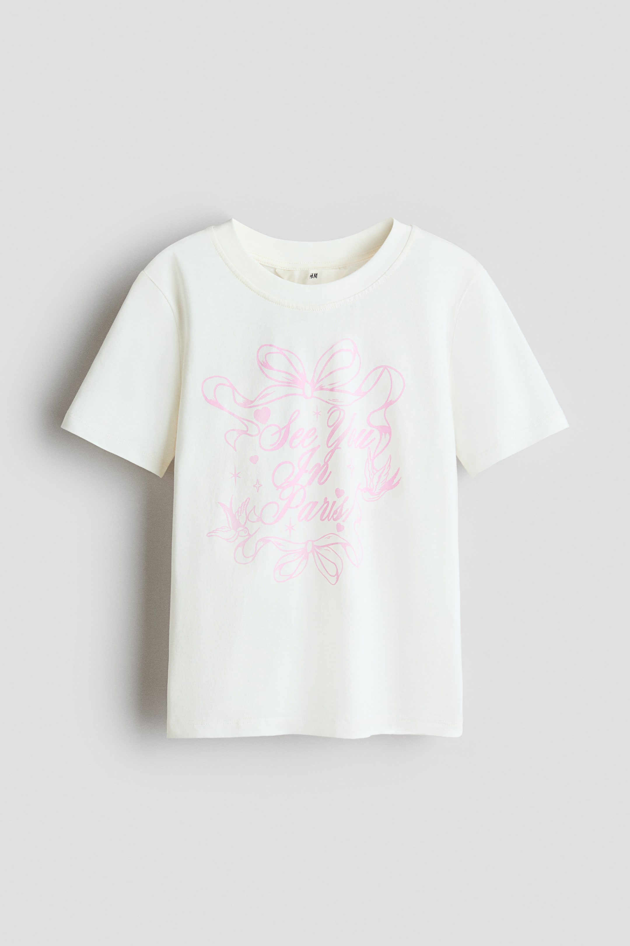 Grösseres Bild ansehen: Ein cremefarbenes T-Shirt zeigt seine Vorderansicht, mit einem runden Halsausschnitt und geripptem Abschluss. Ein rosa Druckmotiv schmückt die Vorderseite, mit dem Text "See You In Paris", umgeben von Schleifen, Herzen und Sternen.
