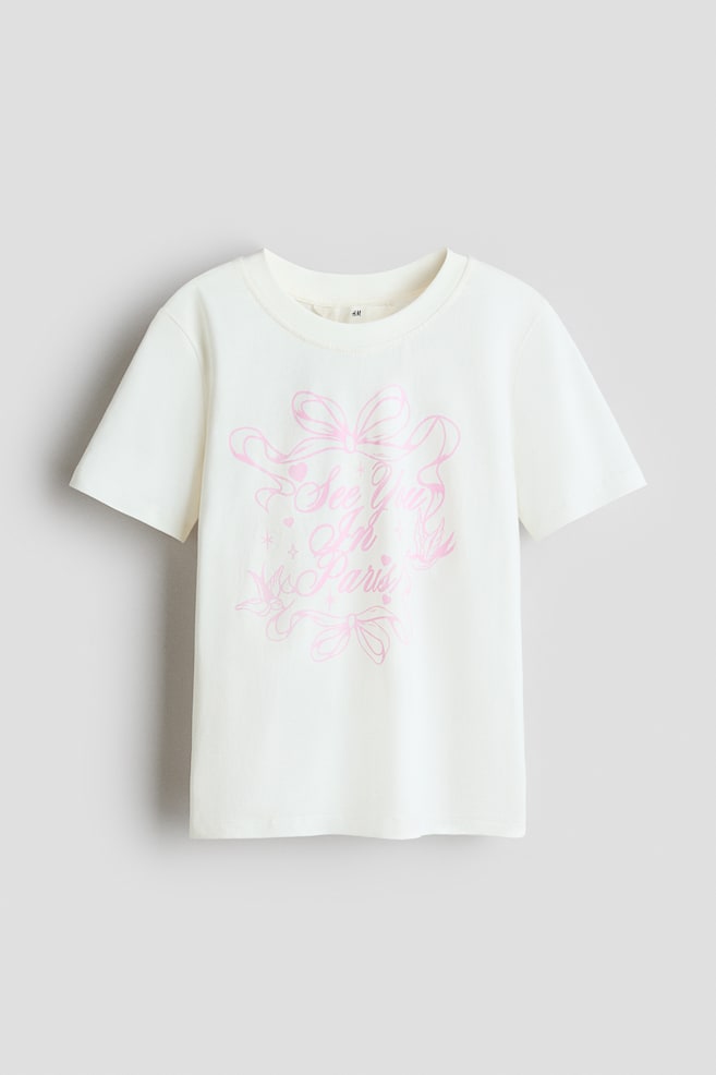A cream t-shirt is presenting its front view, featuring a round neckline with ribbed trim. A pink print motif is decorating the front, displaying "See You In Paris" text surrounded by bows, hearts, and stars. - 1